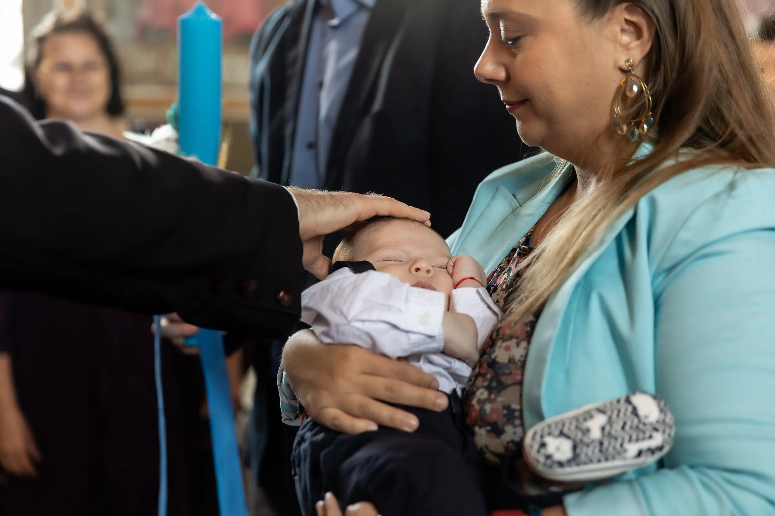 A woman holding a sleeping baby during a religious or ceremonial event, with a man placing his hand on the baby's head and a woman smiling in the background.