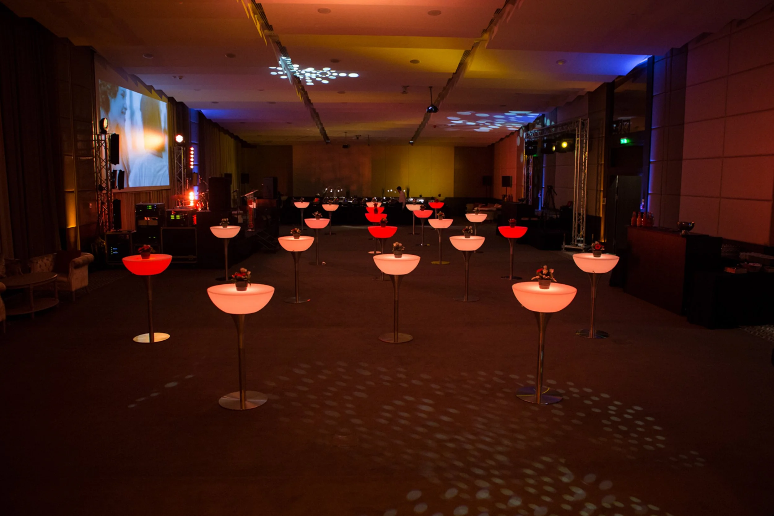 An empty event hall decorated with pink and red flower arrangements on tall stands, with a DJ booth and lighting equipment at the far end, and a large projection screen on the left wall displaying a video or image of a person.