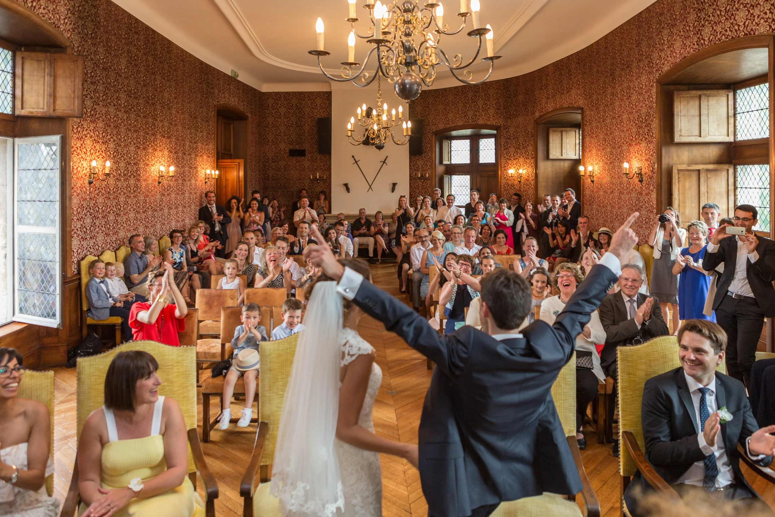 A wedding reception with many guests seated in a decorated room, and a bride and groom dancing in the foreground.
