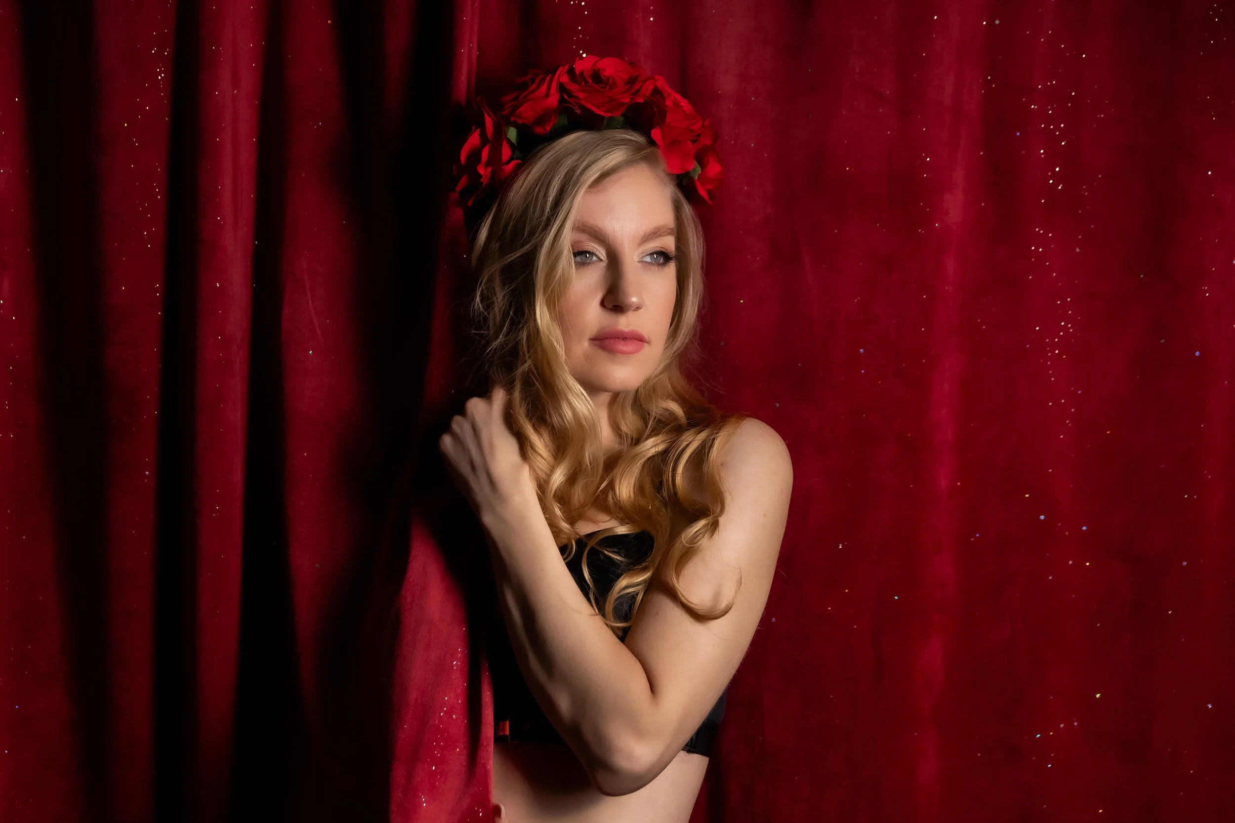 A woman with long blonde hair wearing a black top, standing against a red curtain backdrop with glittering details, and a large red floral headpiece.