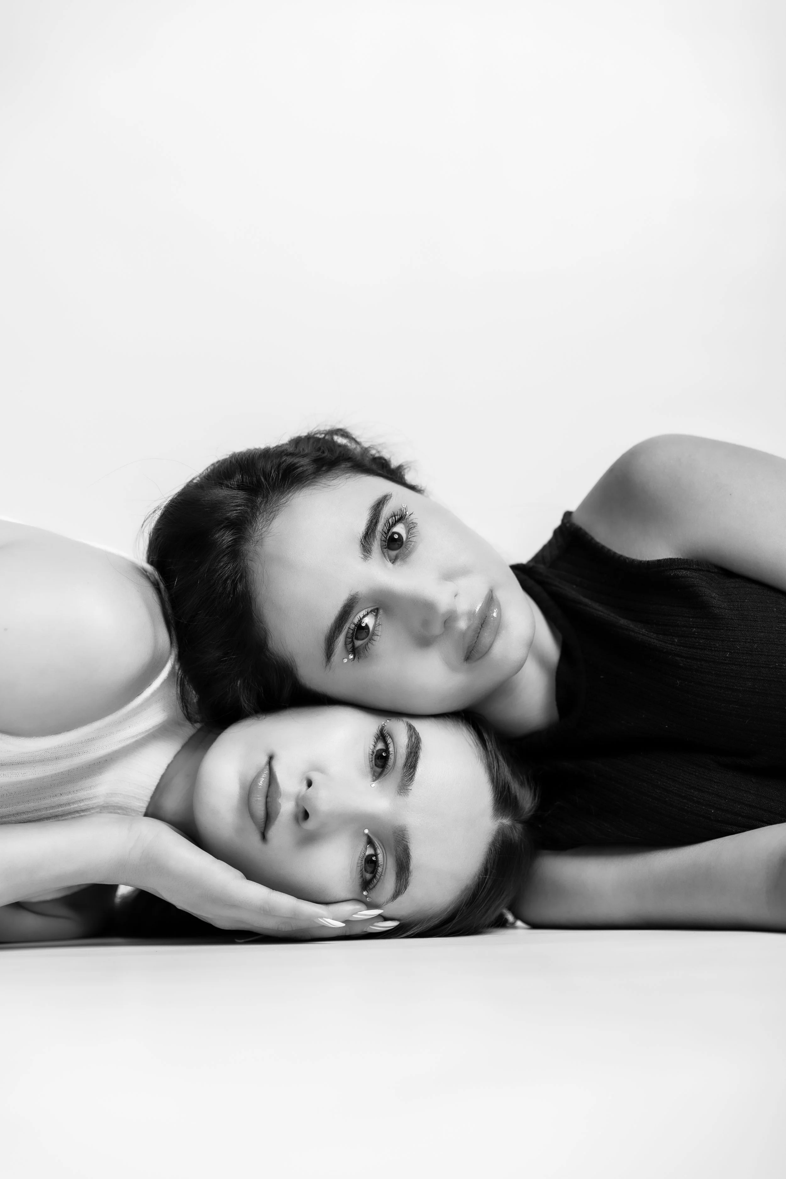 Black and white photo of three women lying close together, with their heads touching. Two women are looking directly at the camera, while the third woman is looking slightly to the side. All have makeup and stylish hair.