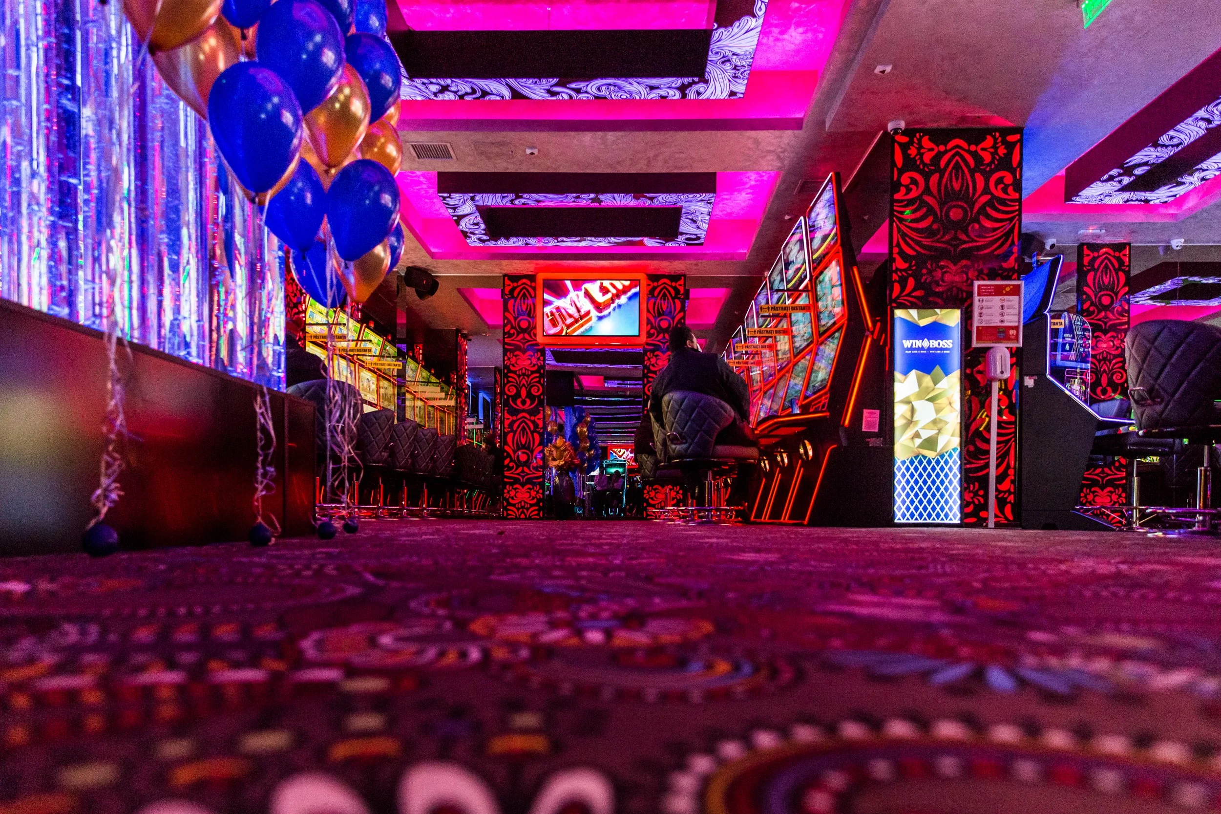 Inside a colorful slot machine casino, with bright LED screens, neon signs, and decorative patterns on the walls, featuring a person sitting in front of slot machines.