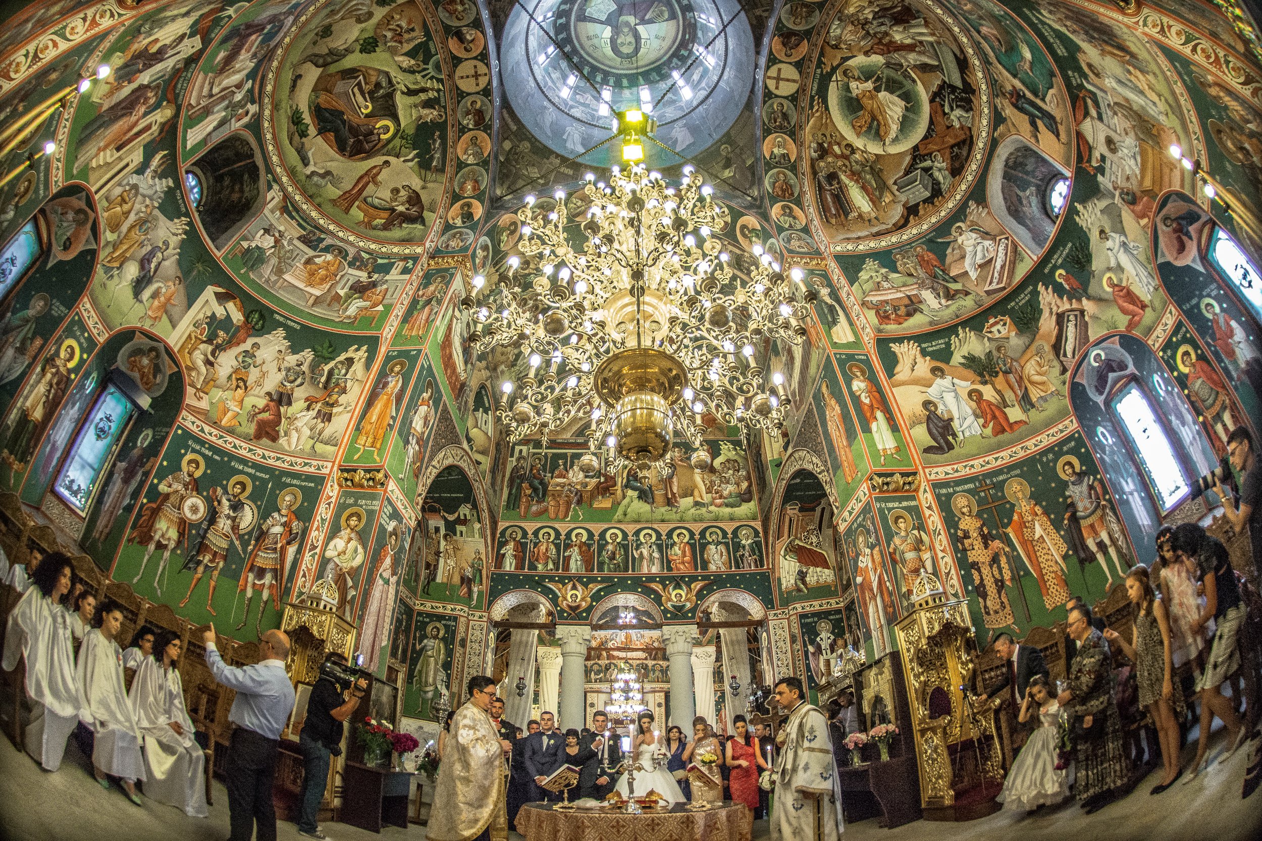 Interior of an ornate church with colorful religious frescoes on the walls and ceiling, a large chandelier hanging in the center, and people gathered for a religious ceremony.