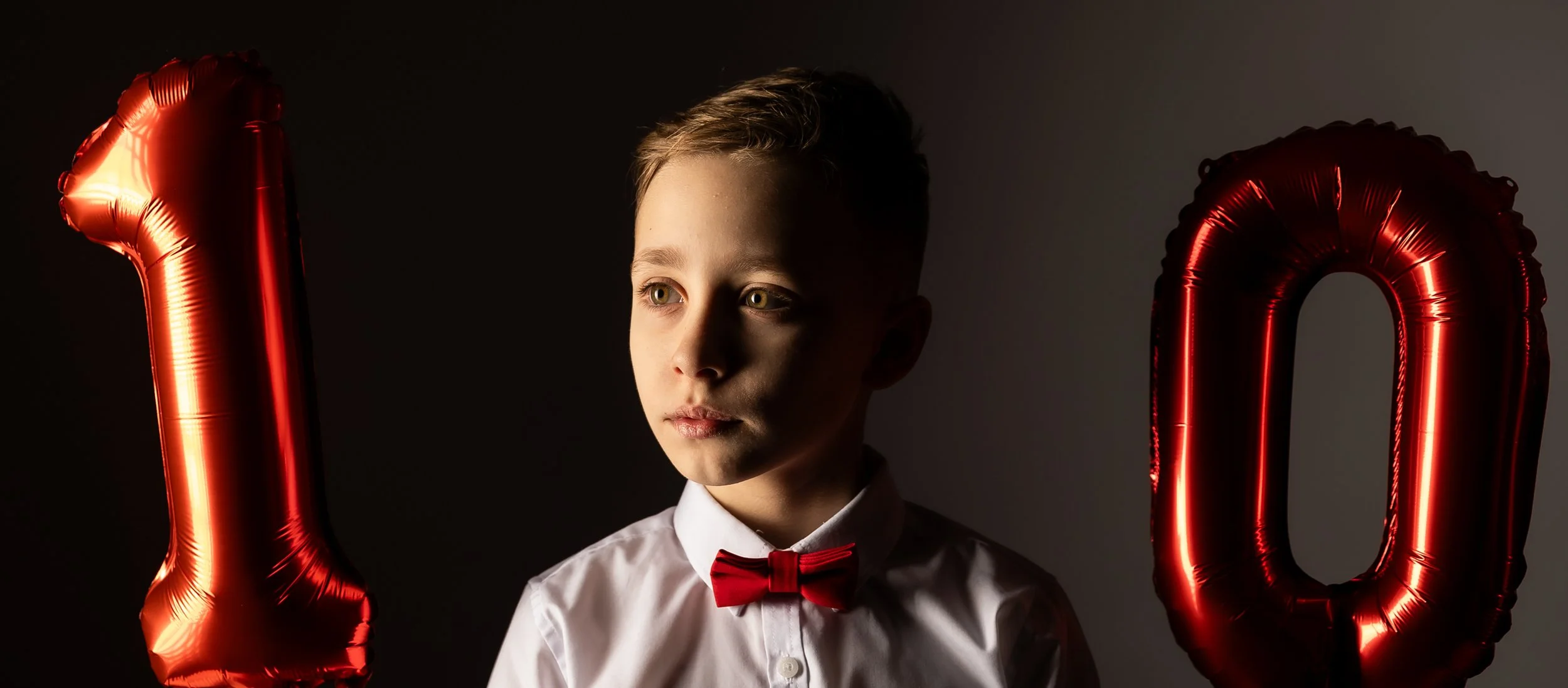 A young boy with light brown hair and green eyes dressed in a white shirt and red bow tie, holding red foil balloons shaped as the number zero, against a dark background.