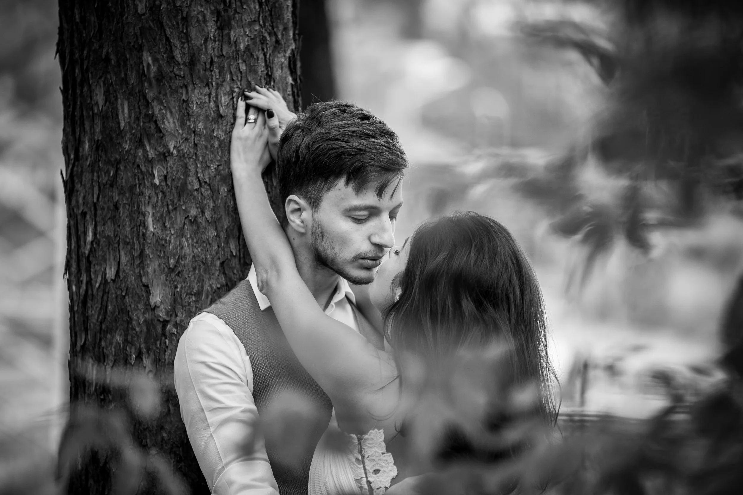 A black and white photo of a young man with short hair and a beard and a young woman with long dark hair, standing close to each other behind a tree. The woman has her arms around the man's neck, and they are looking at each other intimately.