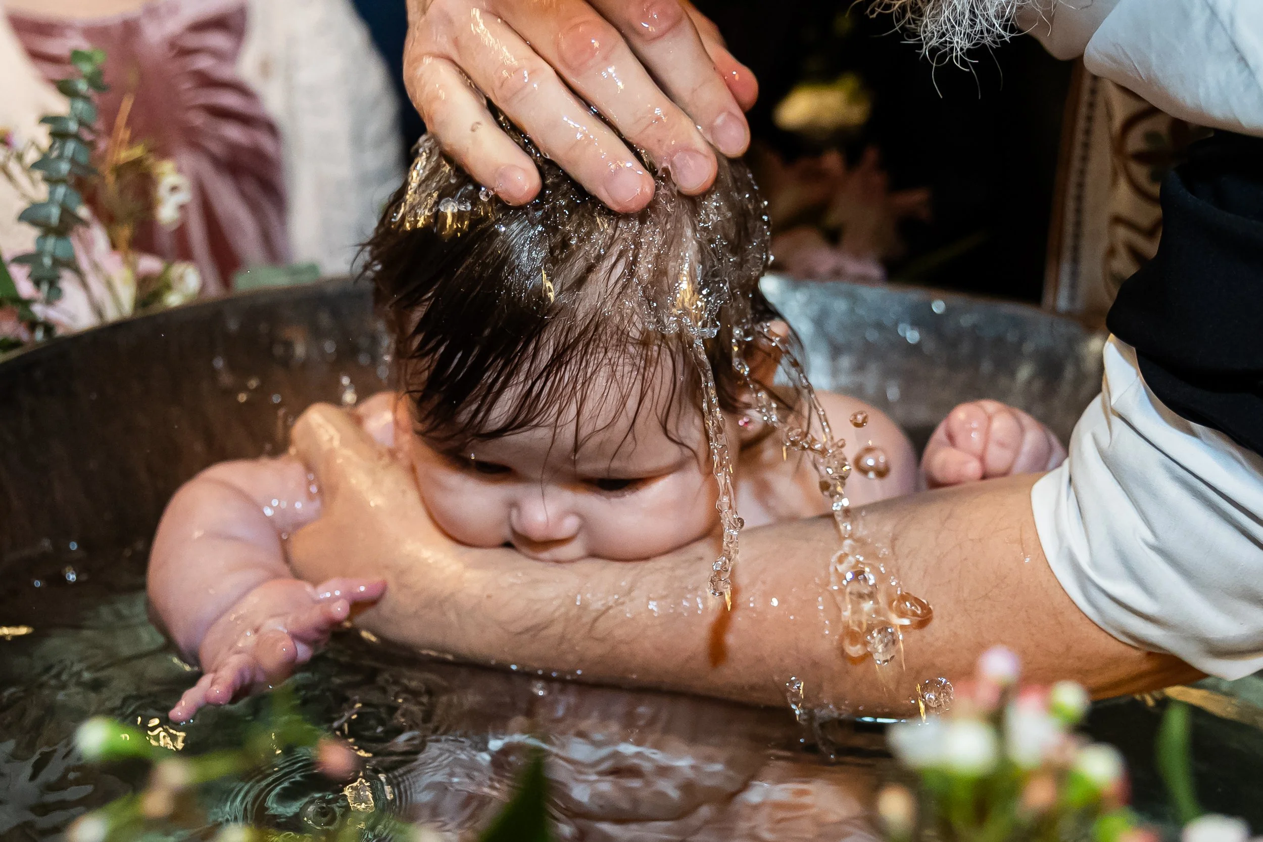 A baby being baptized in a baptismal font with water poured over their head by an adult, with another person nearby