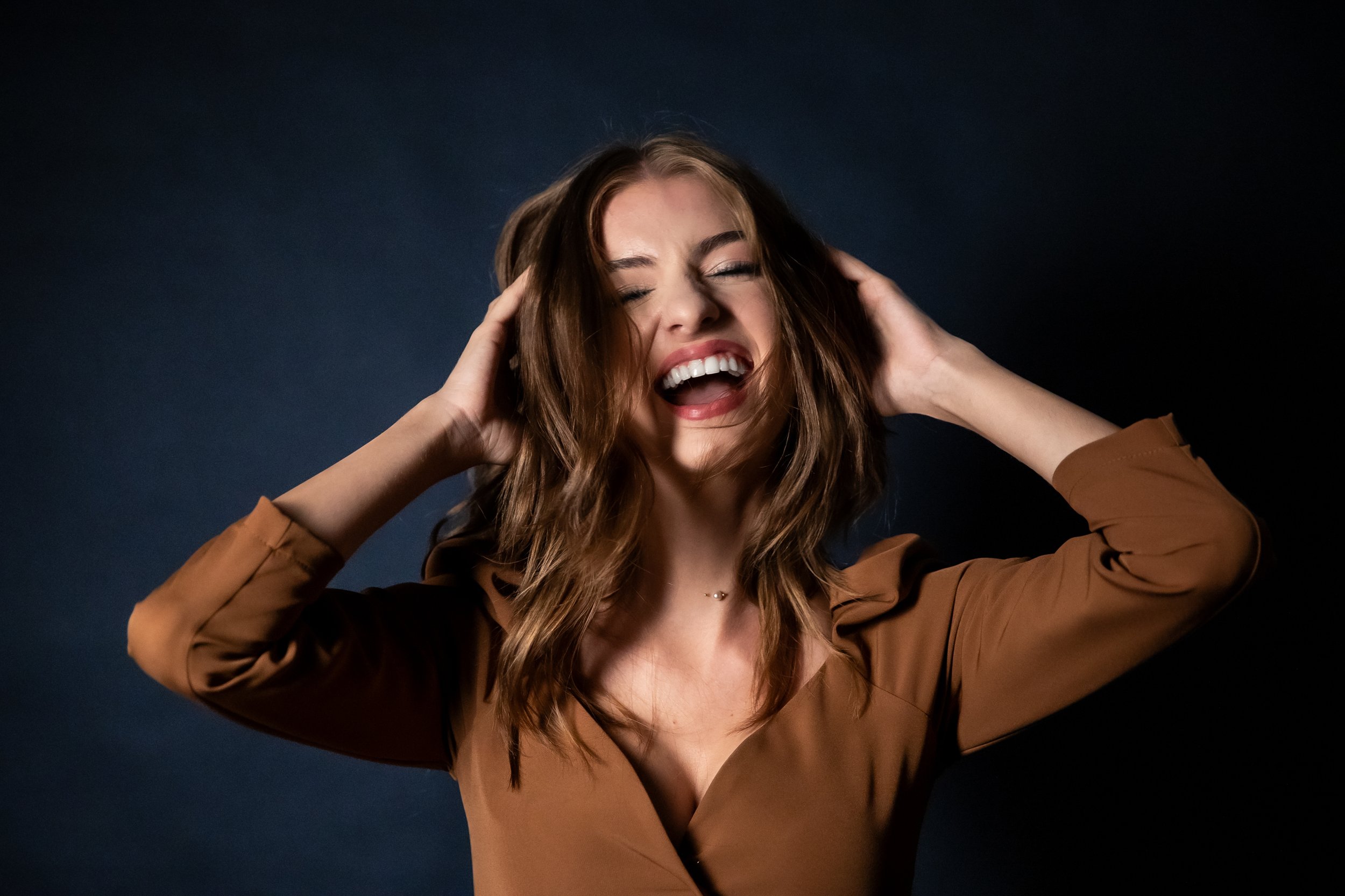 A young woman with long wavy hair smiling and laughing with her eyes closed, holding her head with both hands against a dark background.