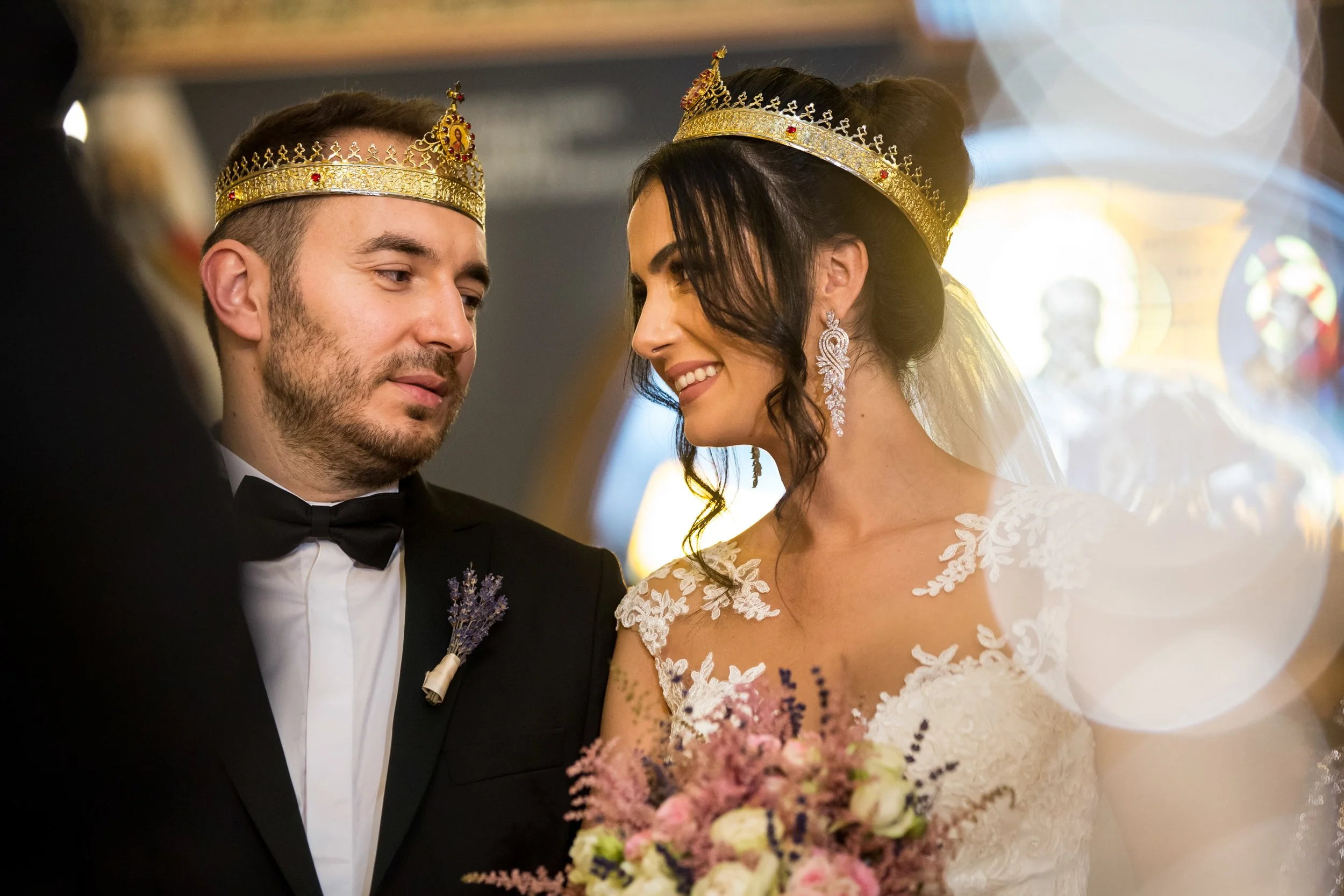 A bride and groom wearing crowns during their wedding ceremony, close-up of their faces, with the bride smiling and holding a bouquet of pink and purple flowers.