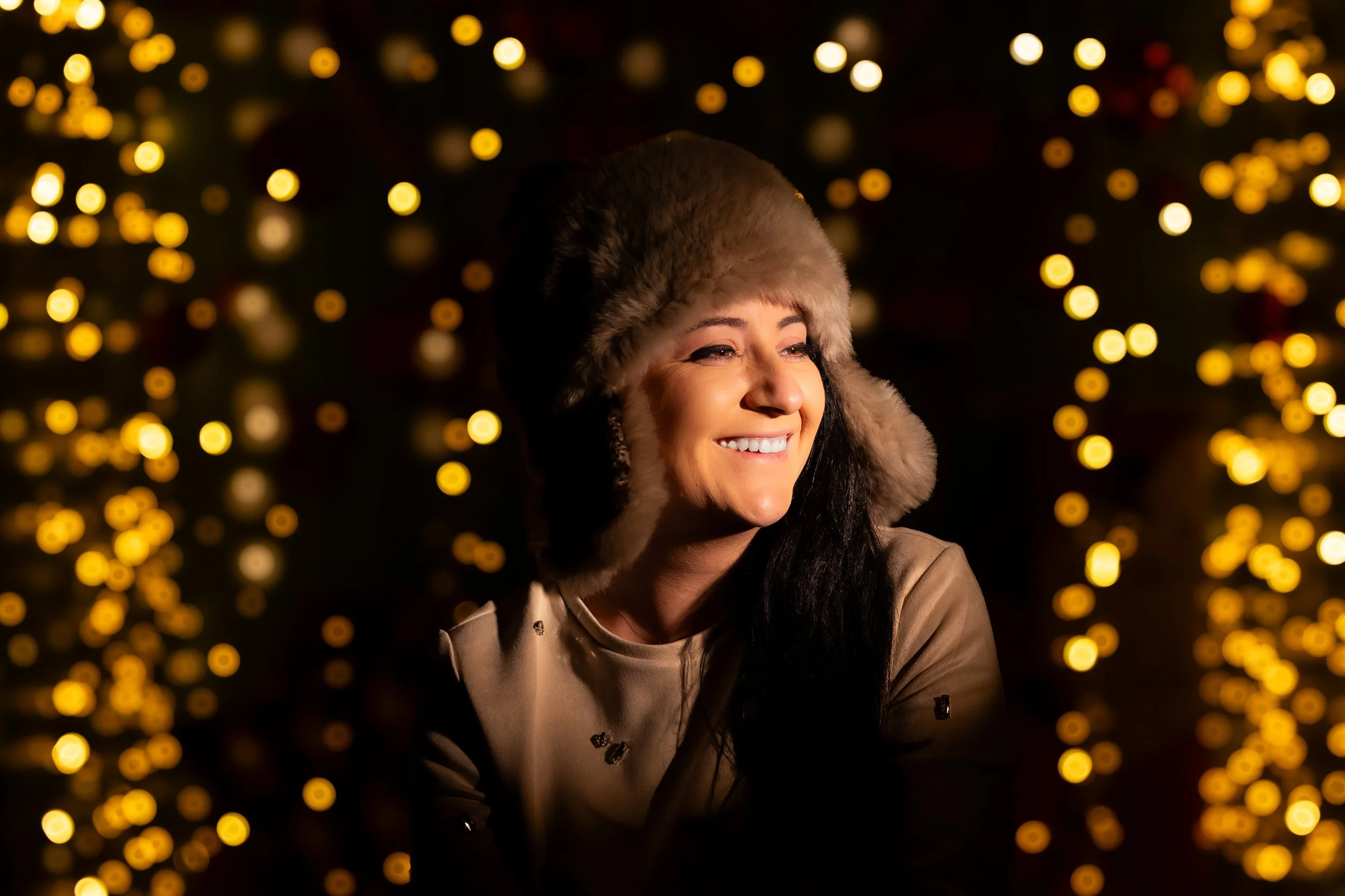 A woman smiling outdoors at night, wearing a furry hat and a tan coat, with blurred yellow lights in the background.