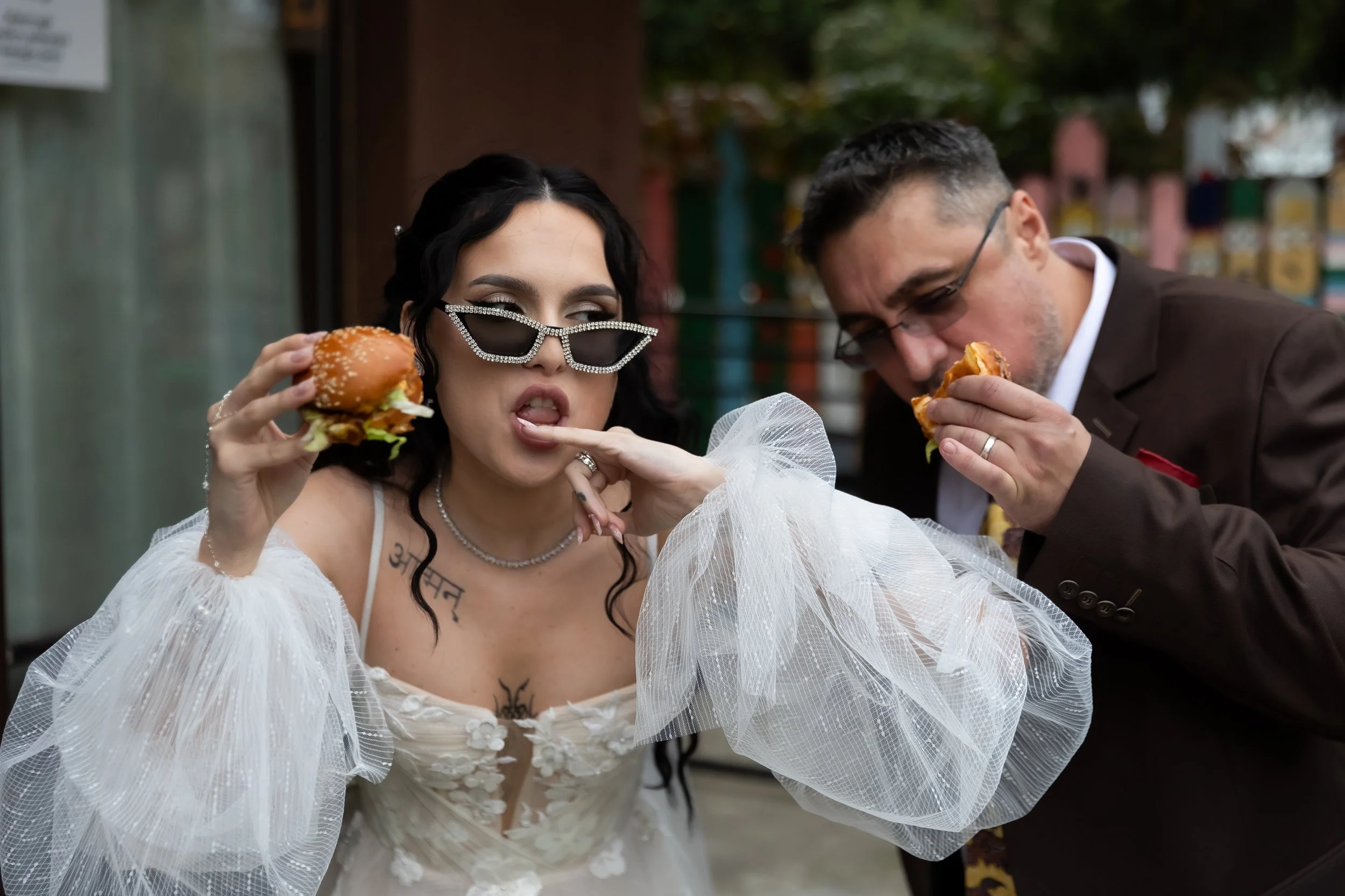 A woman with black hair wearing large sunglasses with rhinestones, a white off-shoulder dress with tulle sleeves, and a man with glasses and short dark hair wearing a brown suit, both eating burgers outdoors.