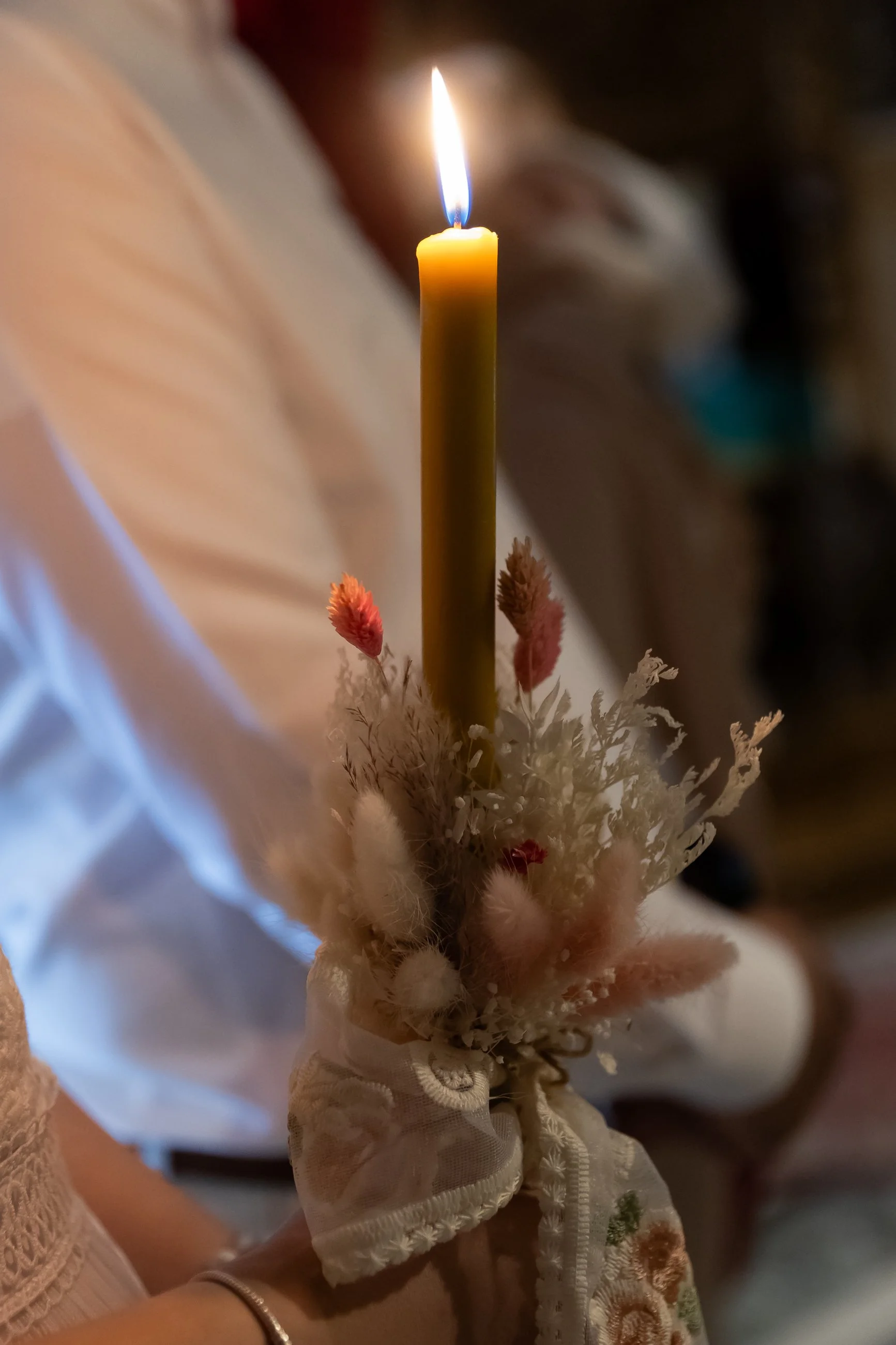 A lit yellow candle held by a person wearing white lace gloves, surrounded by pink and white flowers.