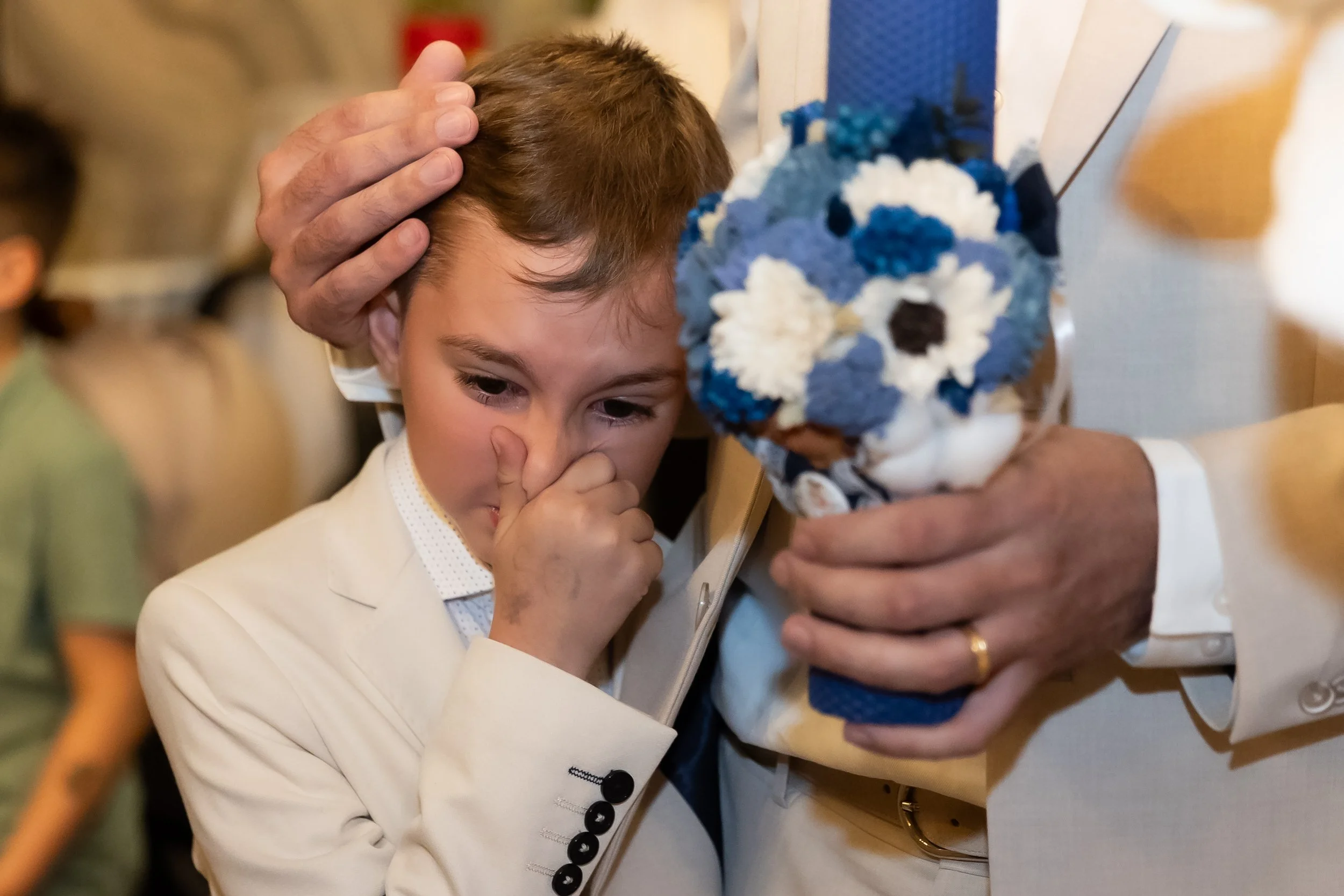 A young boy in a white suit standing at a table, with his hand near his face, looking intently at a blue and white flower arrangement held by an adult. The setting appears to be a celebratory or formal event.