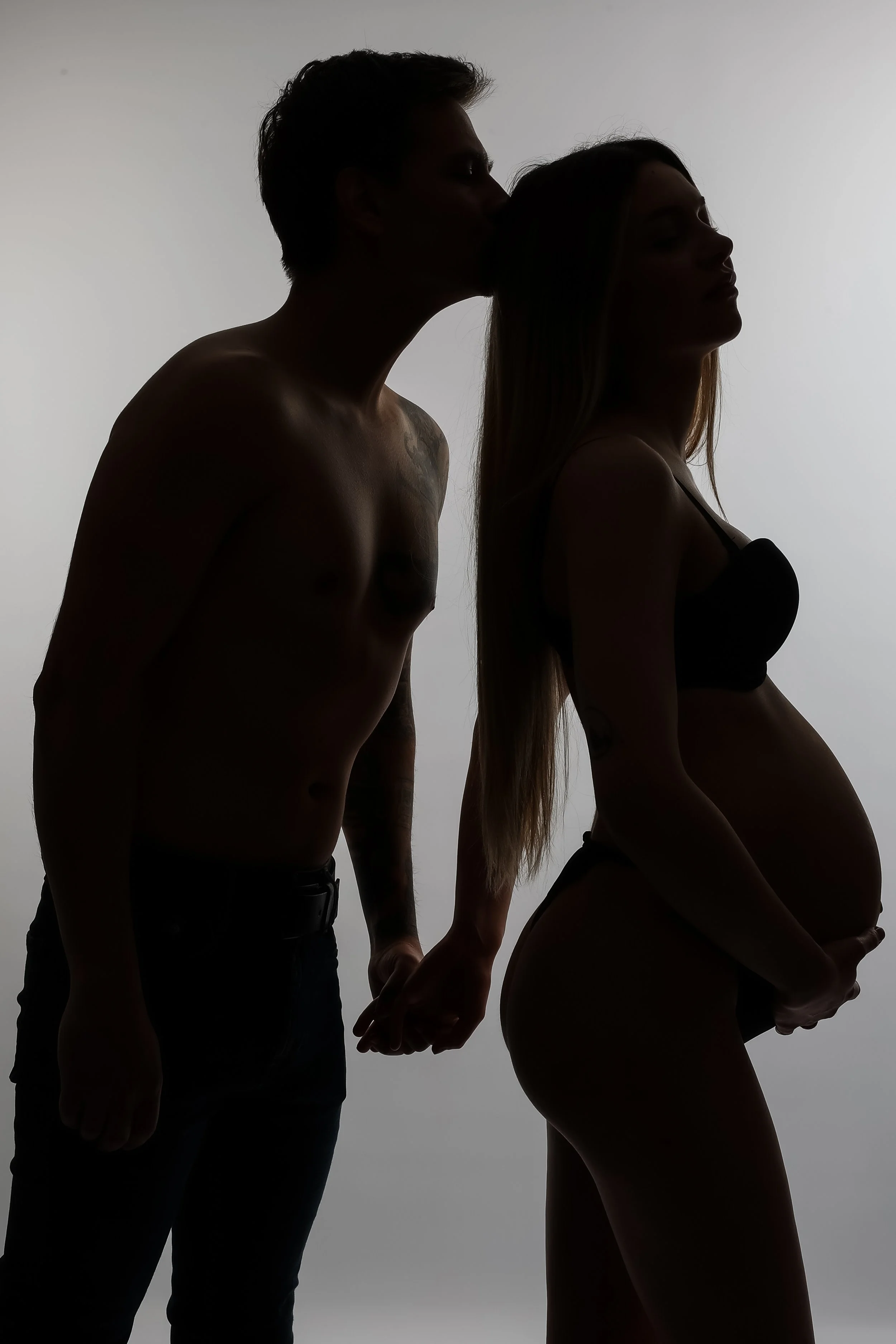 Silhouette of a man and a woman holding hands, with the man kissing the woman's forehead, against a plain background.