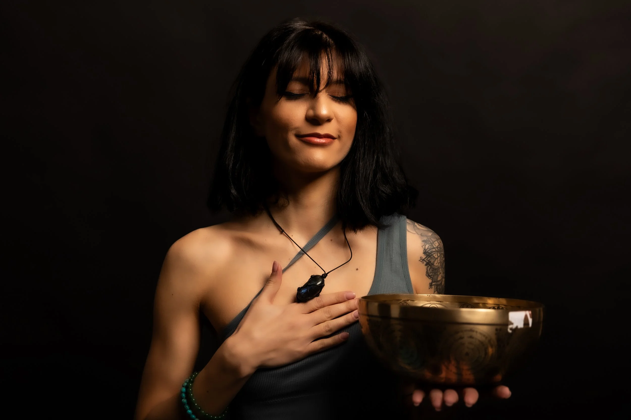 A woman with long black hair, tattoos on her arm, wearing a grey tank top, and a necklace with a large black pendant, is holding a golden bowl and a black stone. She has her eyes closed and appears to be meditating or celebrating a ritual against a d