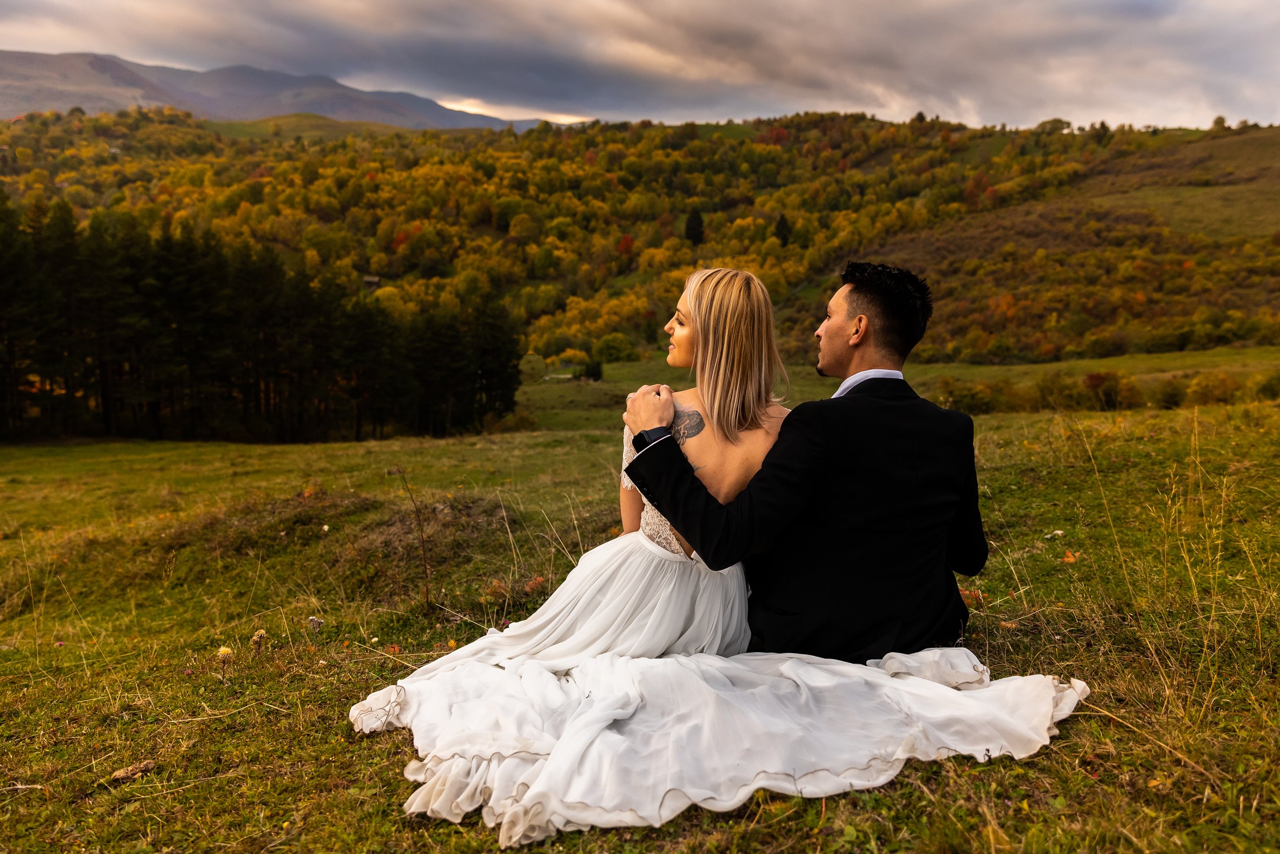 A newlywed couple sitting on grass in a scenic outdoor setting with rolling hills and autumn trees, enjoying a moment together.