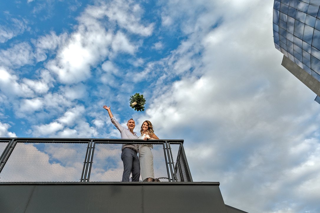 Two people standing on a rooftop balcony, tossing a bouquet into the sky, with blue sky and clouds behind them.