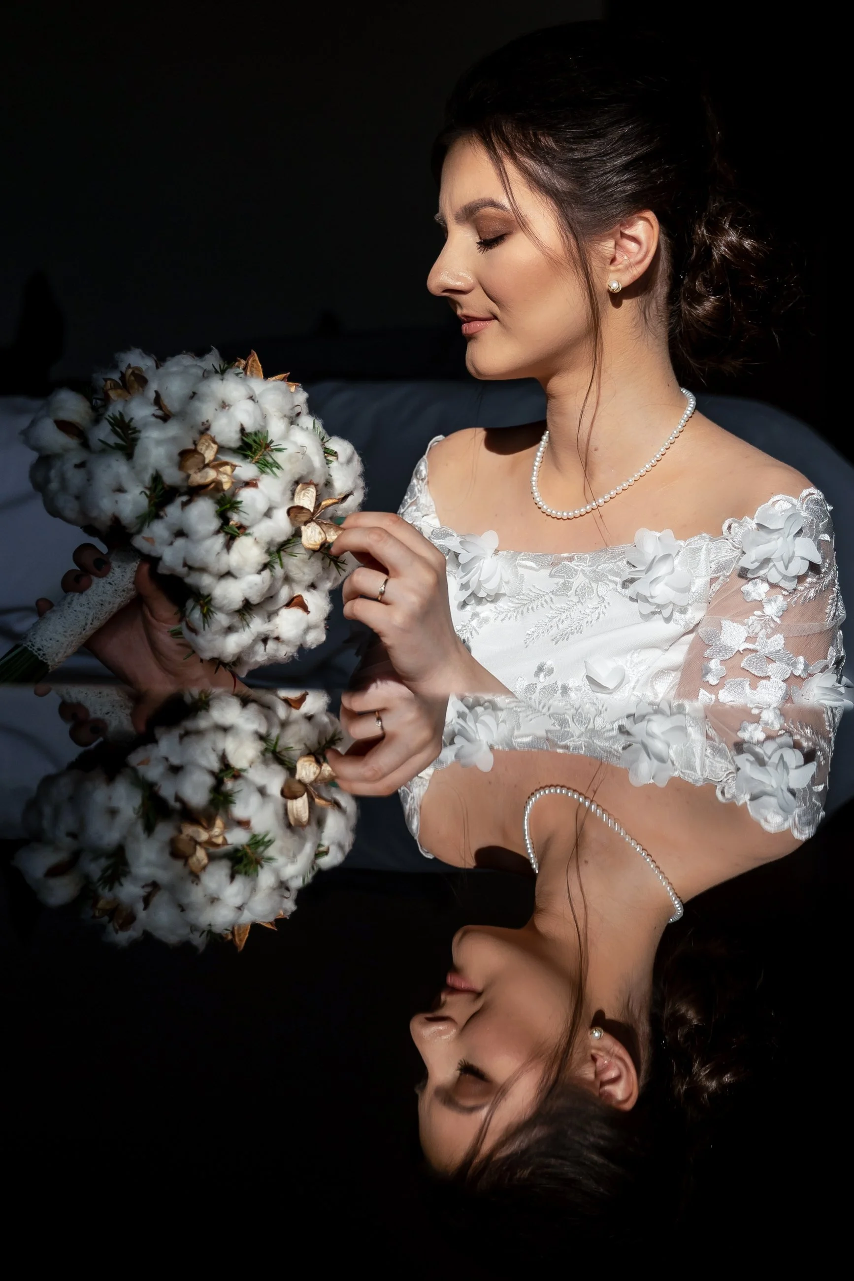 A bride in a white lace wedding dress with floral embellishments, pearl earrings, and a pearl necklace, holding a cotton bouquet with dried leaves and greenery, looking tenderly at the bouquet, against a dark background.