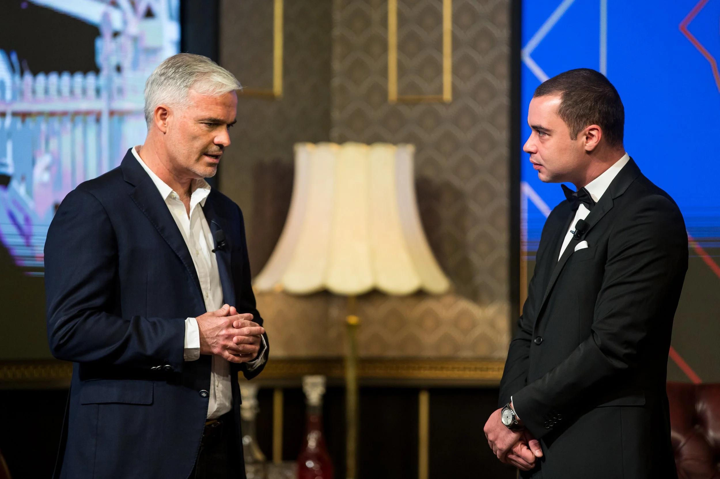 Two men in formal suits with bow ties standing and talking in an elegant room with patterned wallpaper and a lamp in the background.