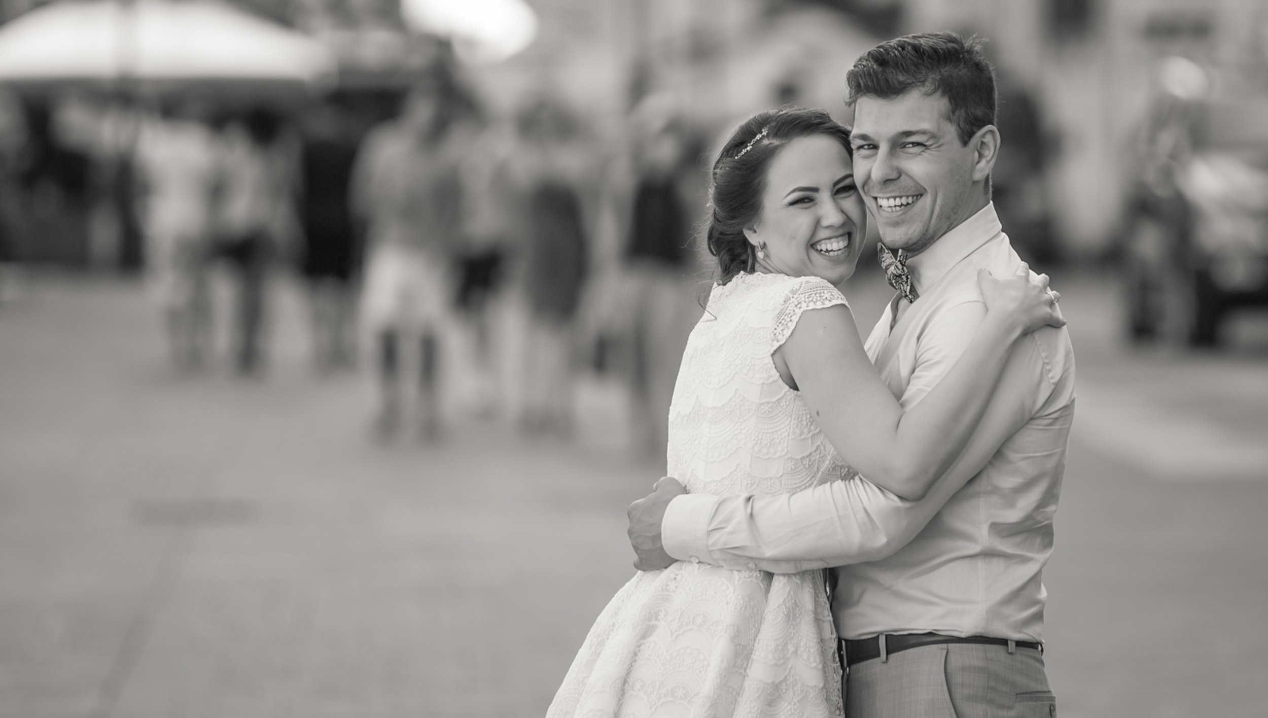 A smiling bride and groom embracing each other outdoors, with a blurred crowd in the background, black and white photo.