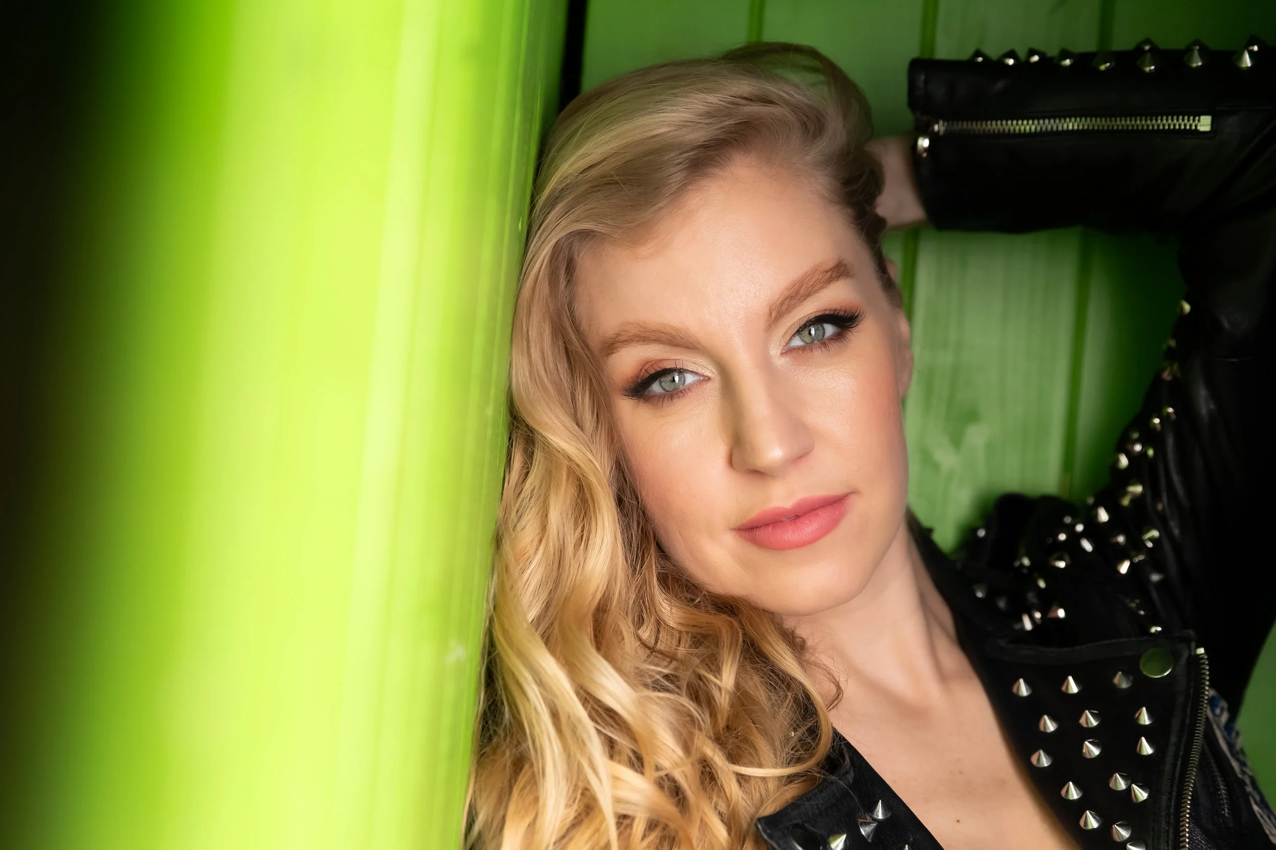 A woman with blonde hair and blue eyes leaning against a lime green wall, wearing a black leather jacket with silver studs.