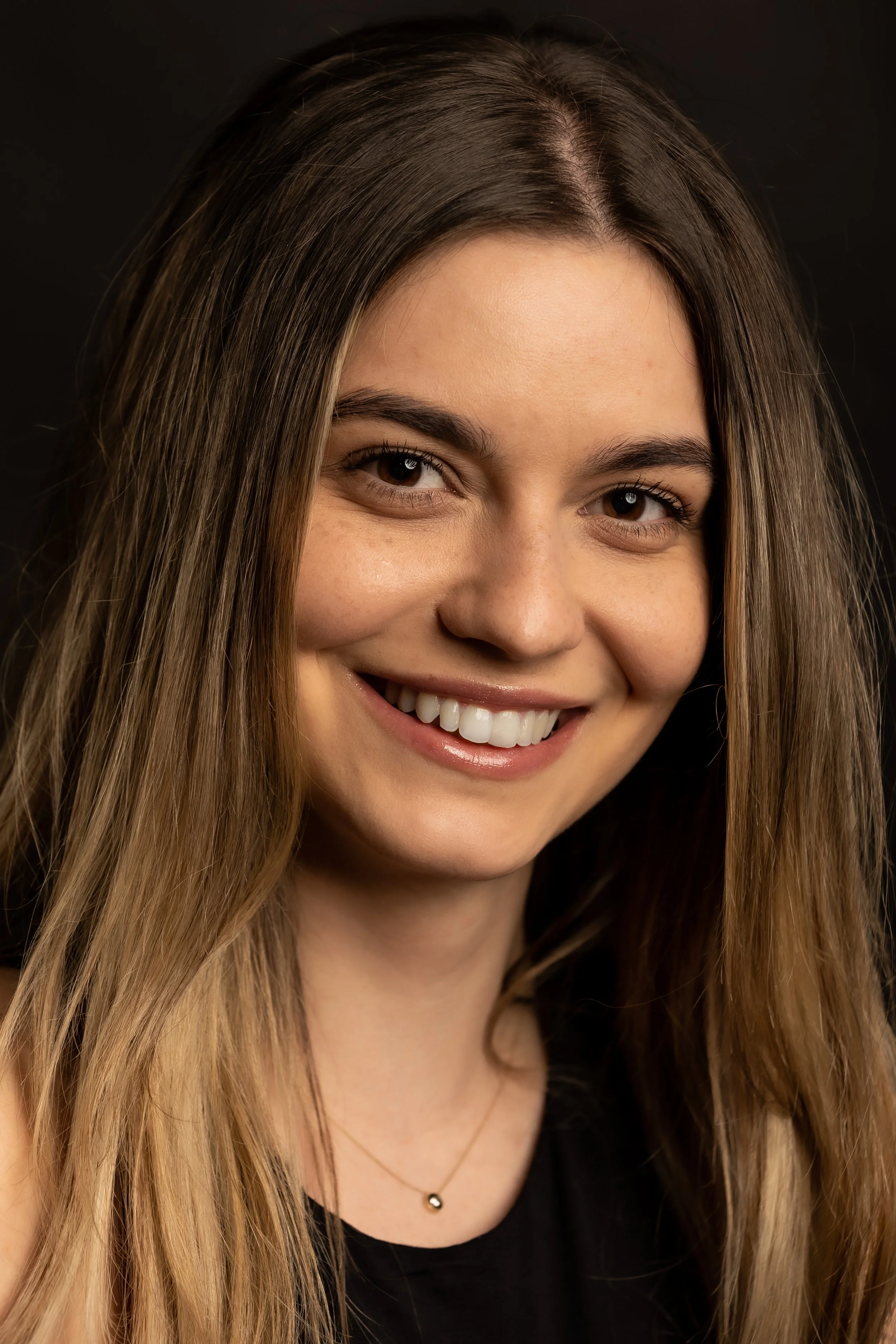 Close-up of a young woman with long, light brown hair, smiling, wearing a black top and a delicate necklace, against a dark background.