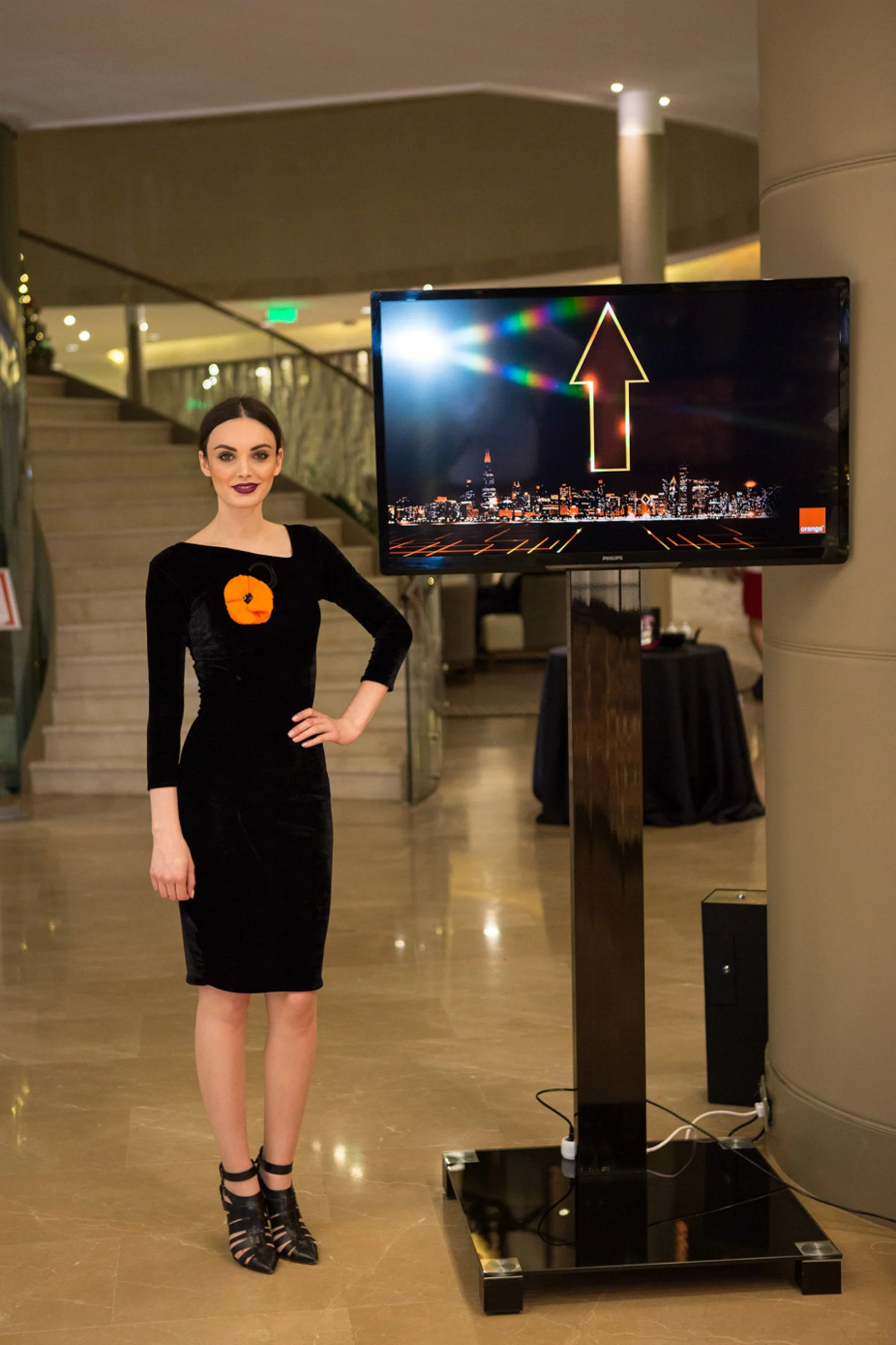 A woman dressed in a black dress with a decorative orange flower pin, standing indoors near a large digital screen displaying a city skyline at night with a glowing arrow pointing upward.