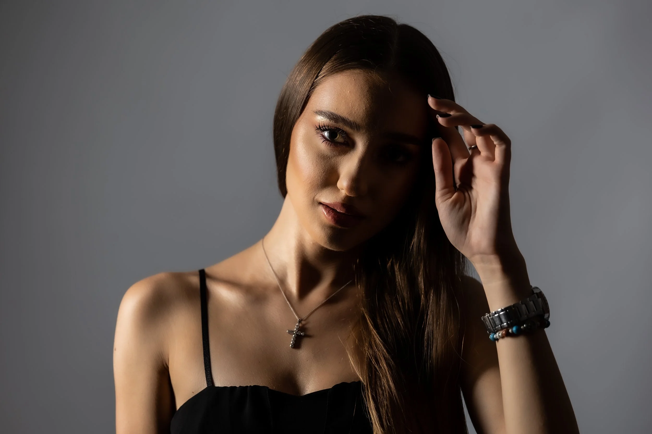 A young woman with long brown hair and light skin, wearing a black tank top, jewelry, and bracelets, looking directly at the camera with her hand near her forehead, against a plain gray background.