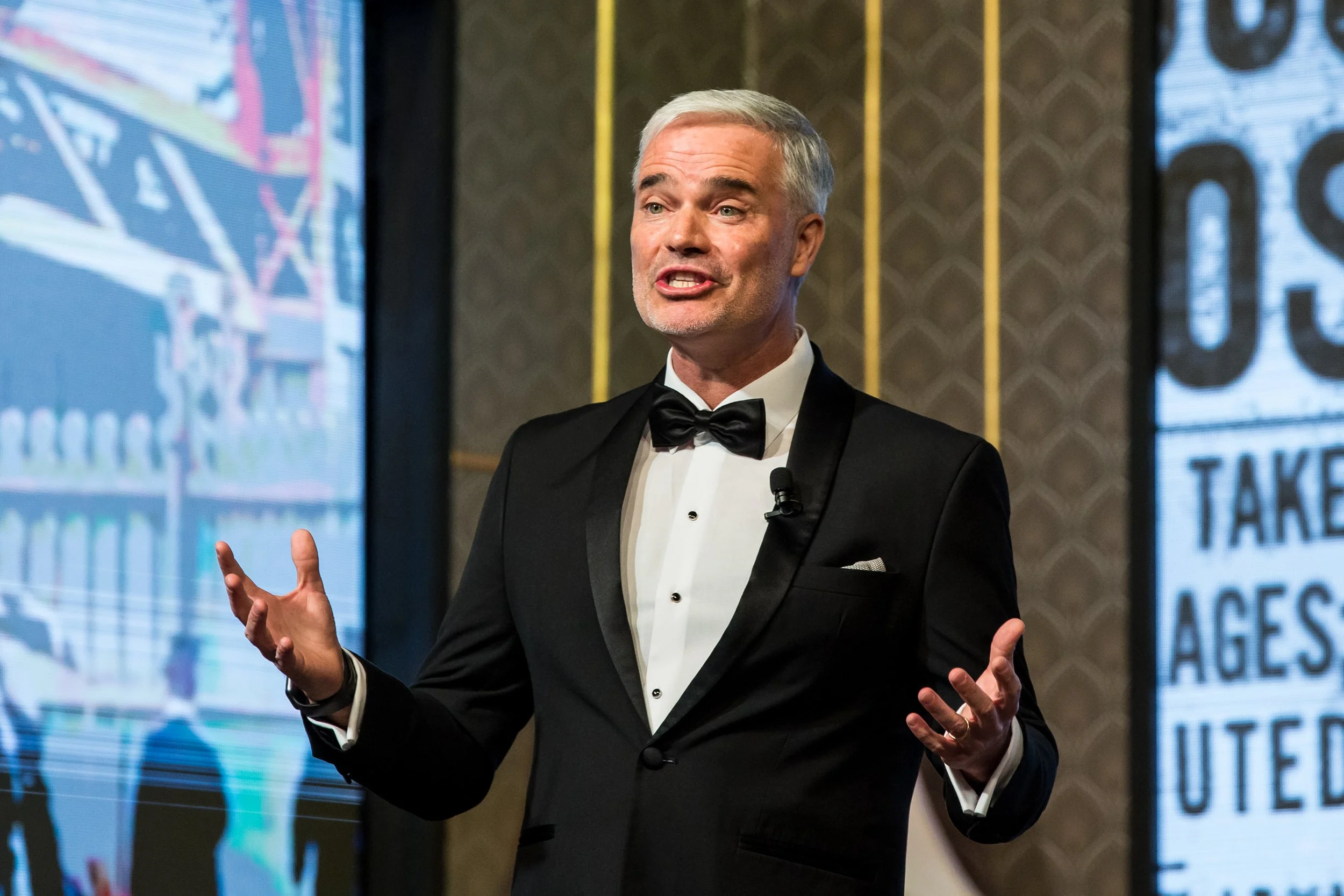 A man with gray hair and a gray beard, dressed in a black tuxedo with a bow tie, speaking at a presentation or event with large screens in the background.