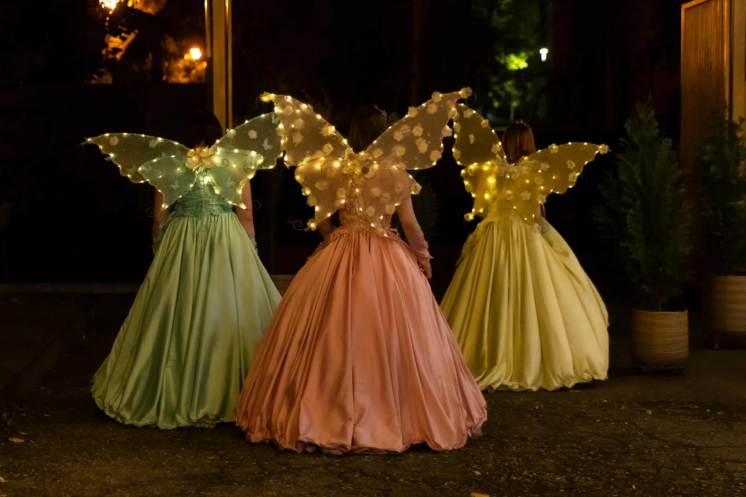 Three women dressed in ball gowns and fairy-themed illuminated butterfly costumes with glowing lights and floral decorations are walking at night outdoors.