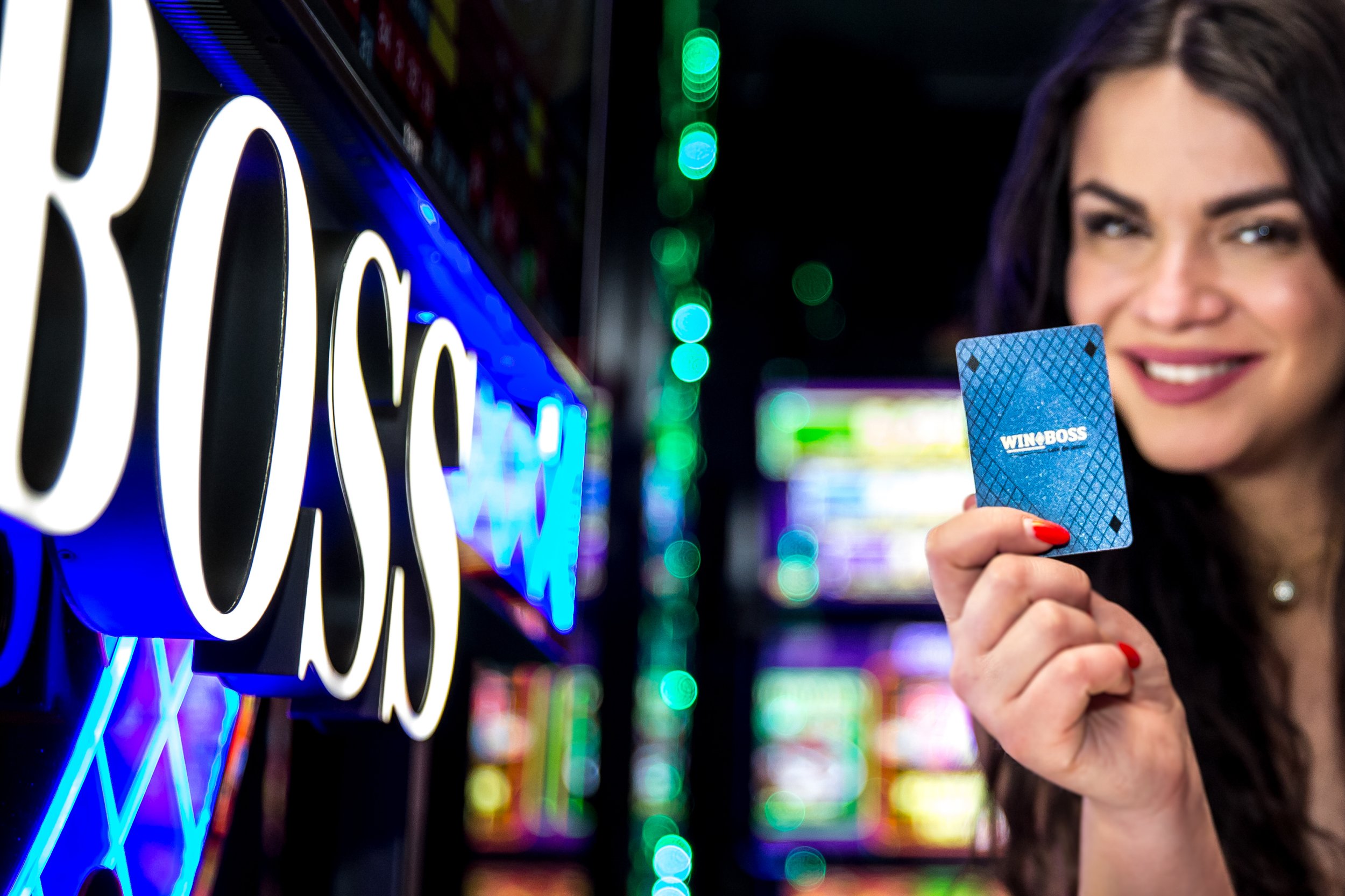 A woman with dark hair and red painted nails holding a blue WynnBOSS gift card in front of illuminated neon signs at an arcade or gaming venue.
