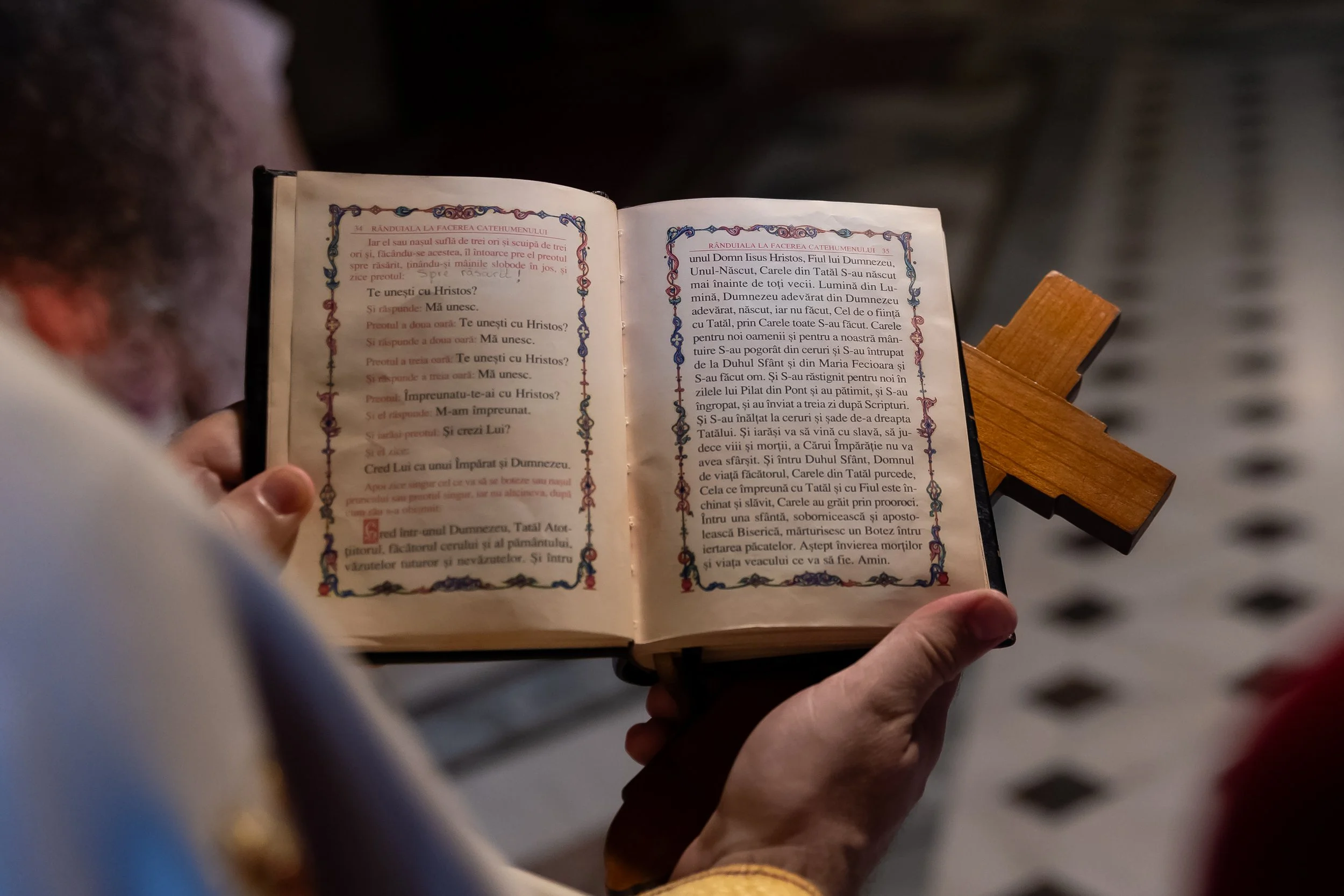 Close-up of a person holding an open religious book with decorative borders, standing on a wooden stand, with a blurred background.