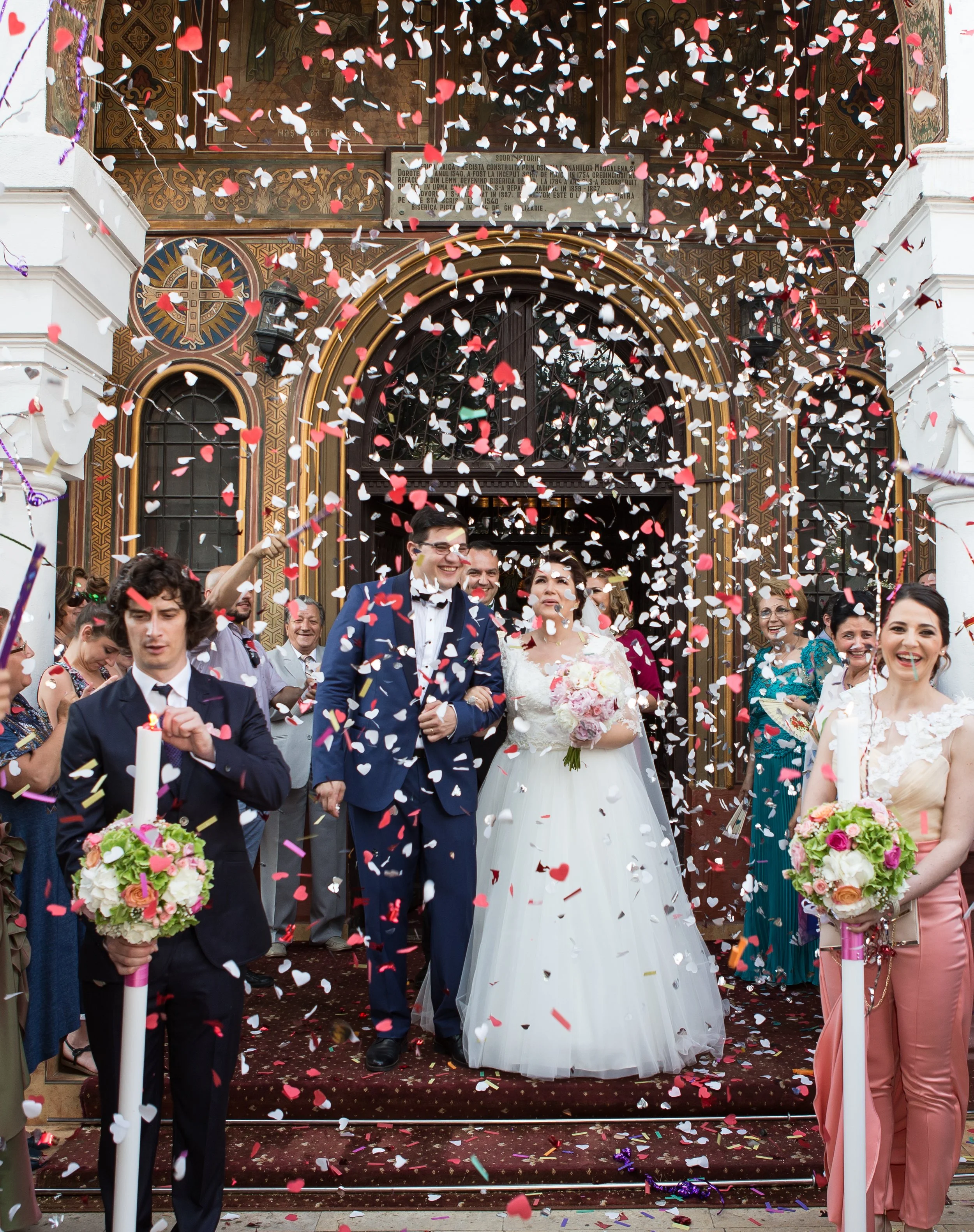 A wedding celebration with a bride and groom walking out of a church surrounded by guests. Colorful confetti is falling from above, and the couple is smiling. Guests hold flowers and candles, and the church background is ornate.