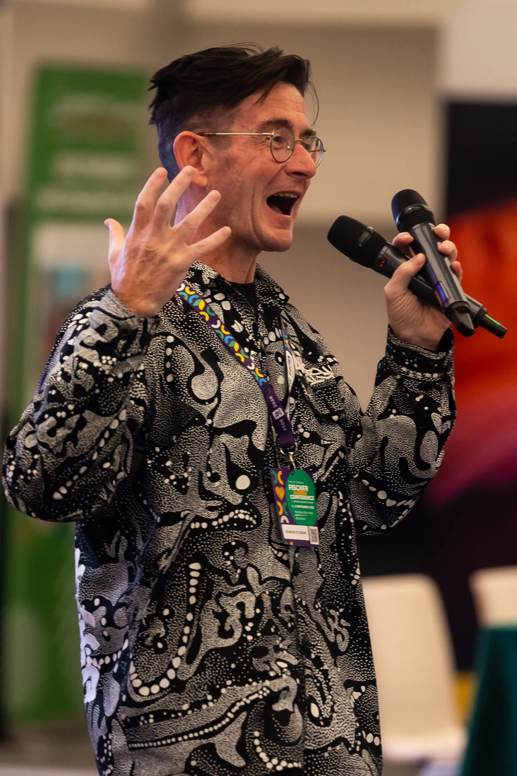 A man with dark hair, glasses, and a patterned black and white shirt holding a microphone and speaking passionately.