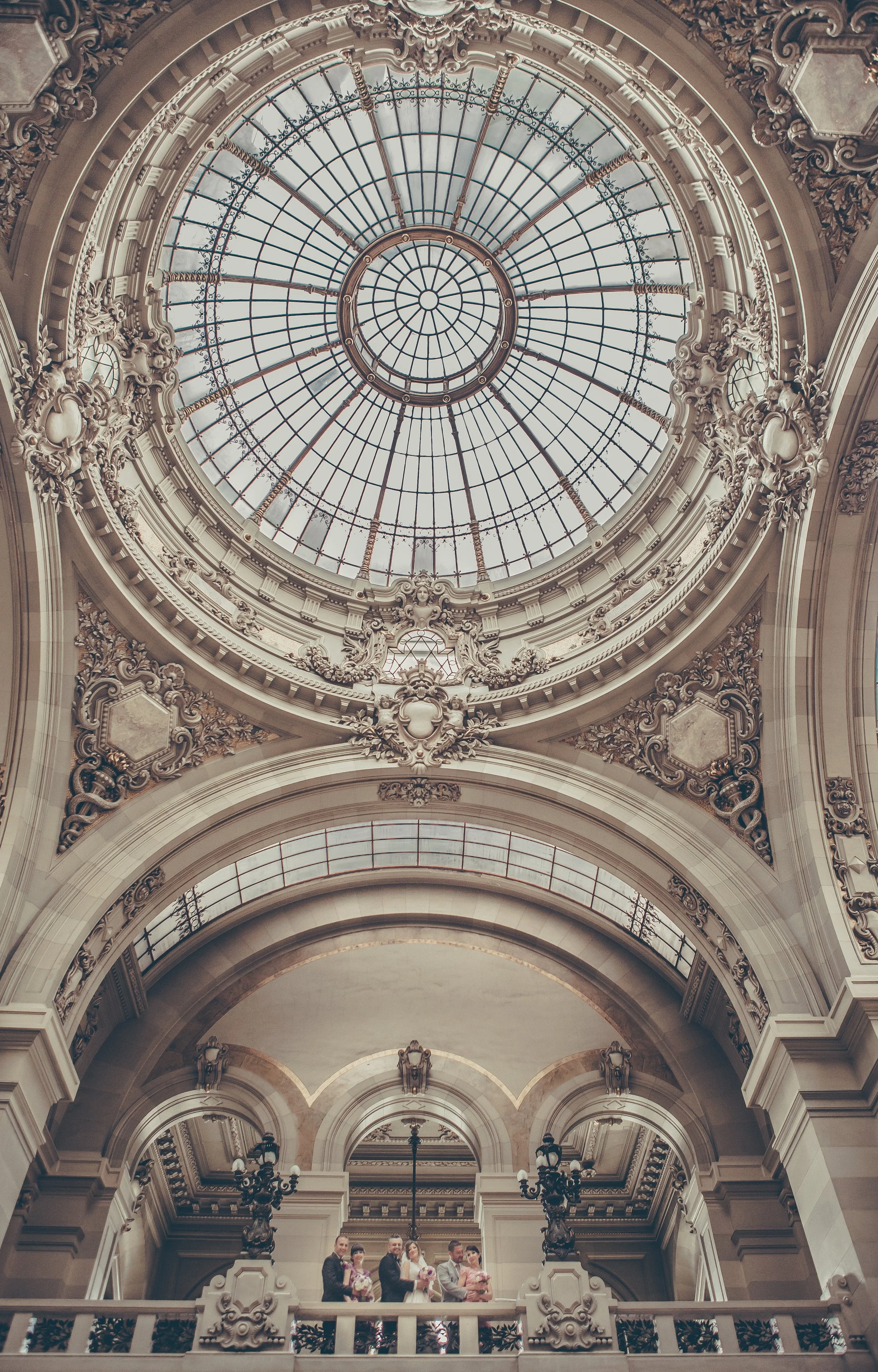 People standing on a balcony inside a grand hall with an ornate glass dome ceiling.