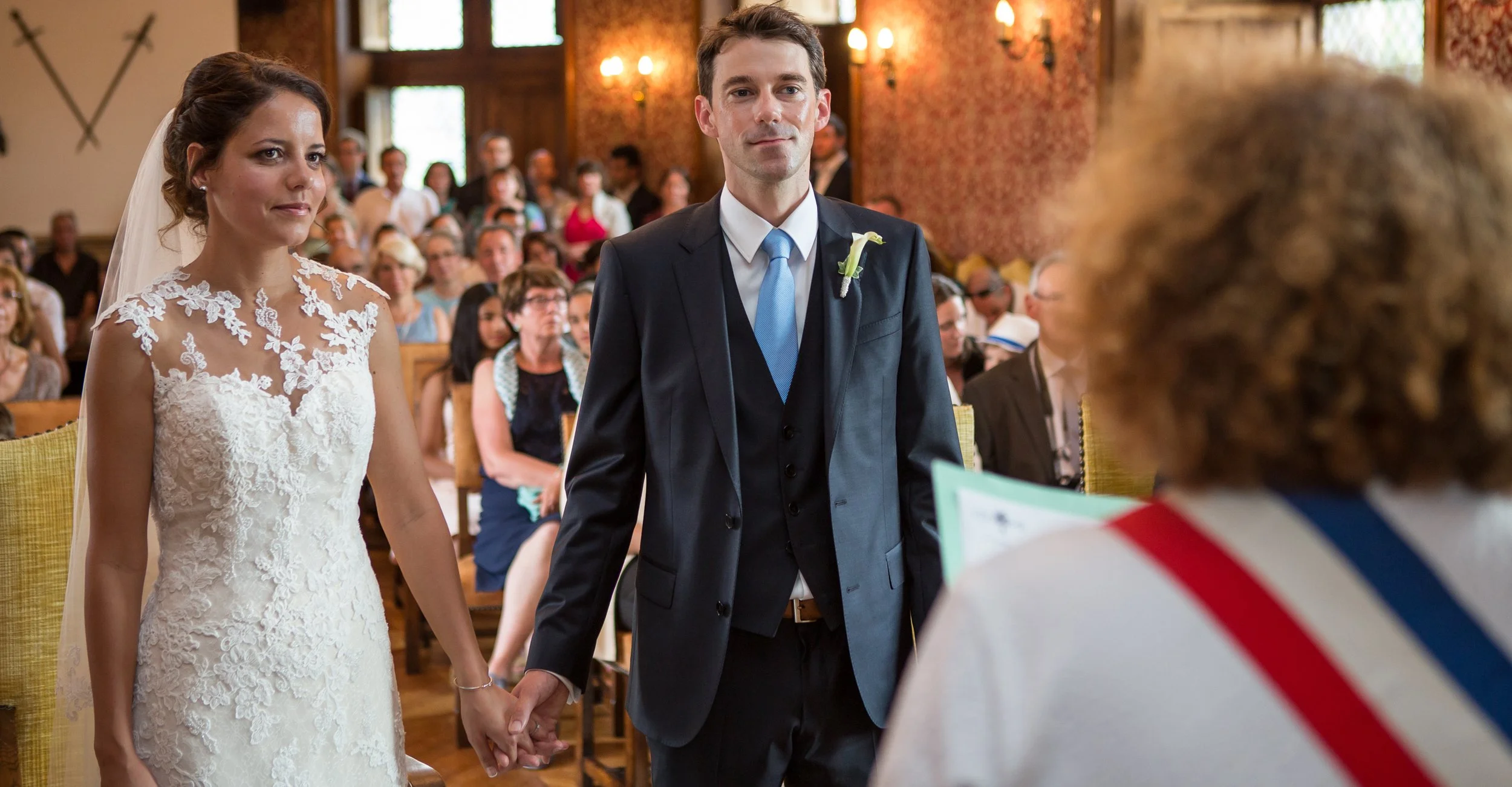A bride and groom hold hands during a wedding ceremony inside a decorated room, with guests seated behind them.