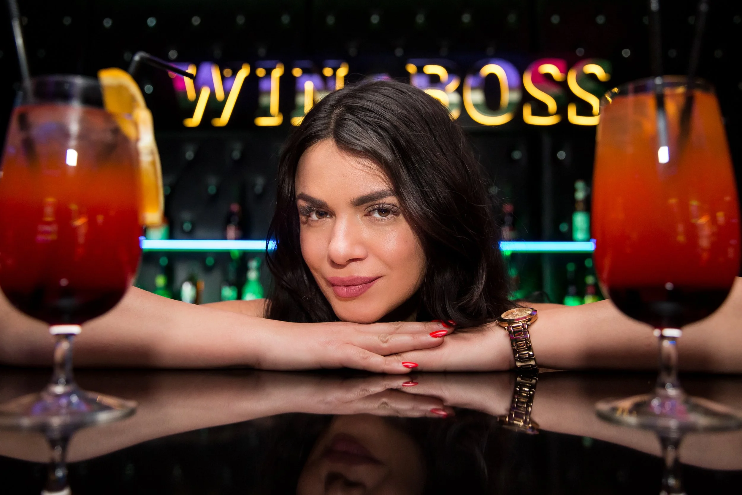 A woman with dark hair and red nail polish resting her arms on a bar counter, with two colorful drinks in glasses in front of her, against a background with illuminated signs.