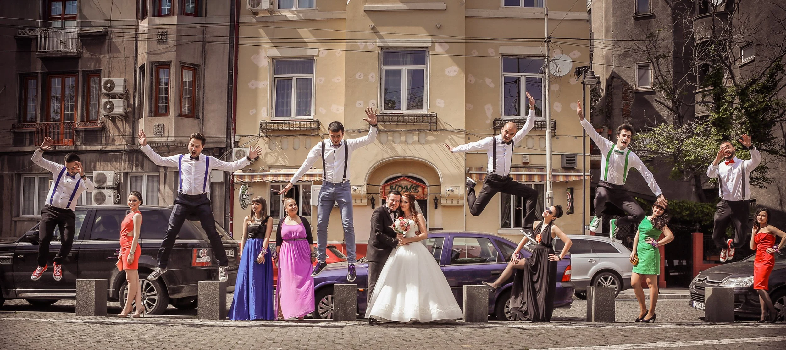 A wedding celebration with the bride and groom in the center, surrounded by friends and wedding party members, some of whom are jumping in the air, in an urban street setting.