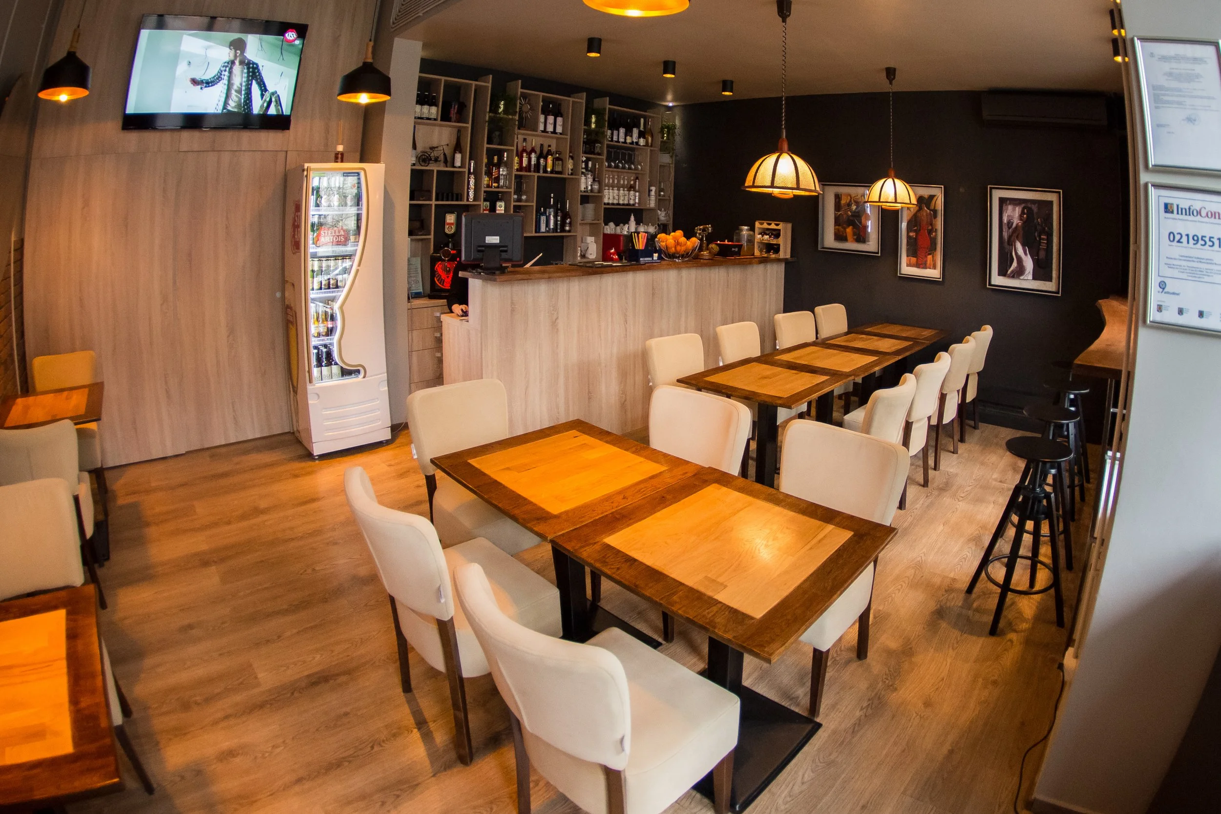 A cozy restaurant interior with wooden tables and cream-colored chairs. The bar area features shelves stocked with bottles and glasses, with framed artwork on a dark accent wall. Pendant lights hang from the ceiling, providing warm lighting. A flat-s