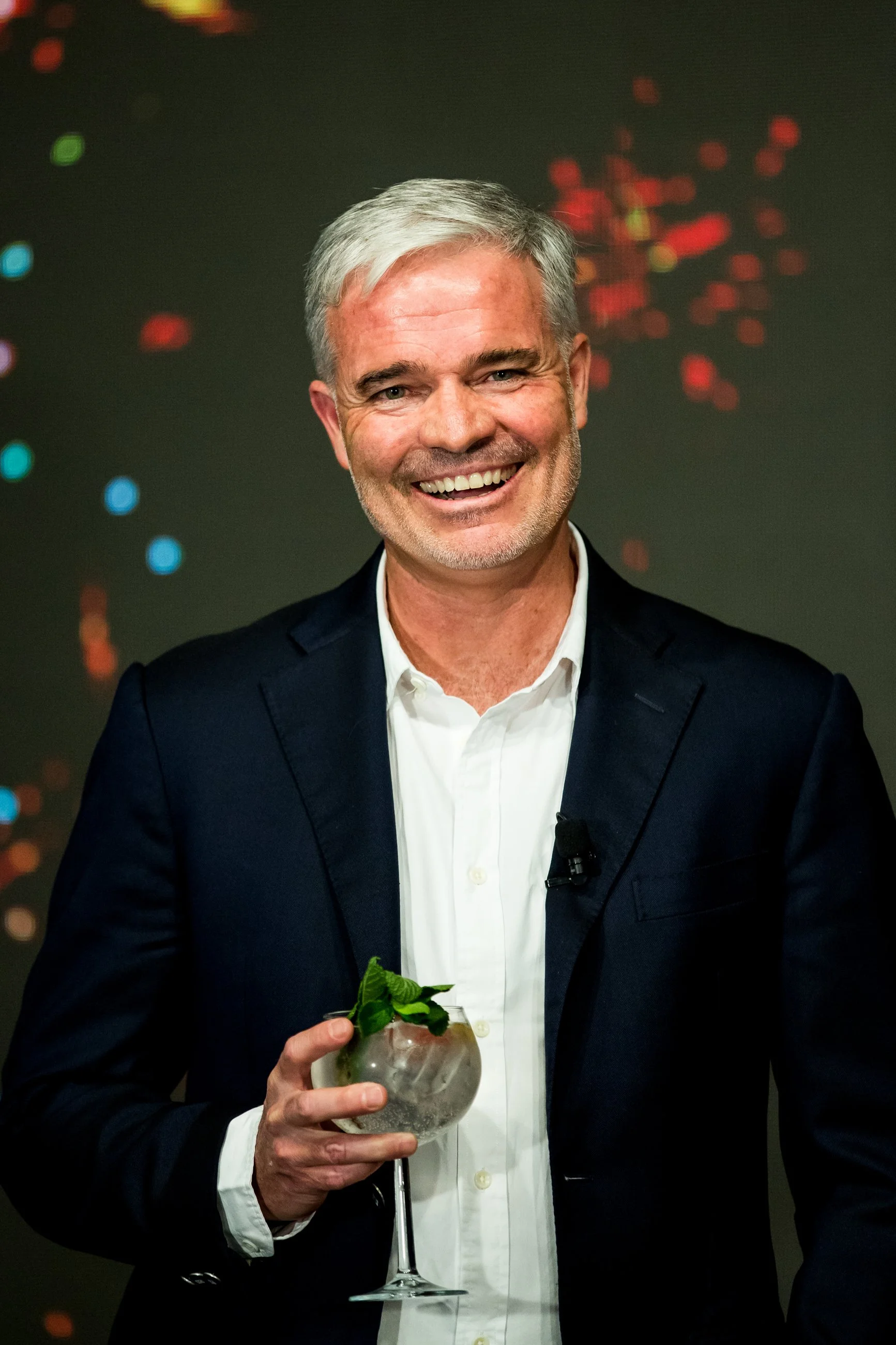 A smiling middle-aged man with gray hair and a beard, dressed in a dark suit and white shirt, holding a cocktail glass with mint leaves.