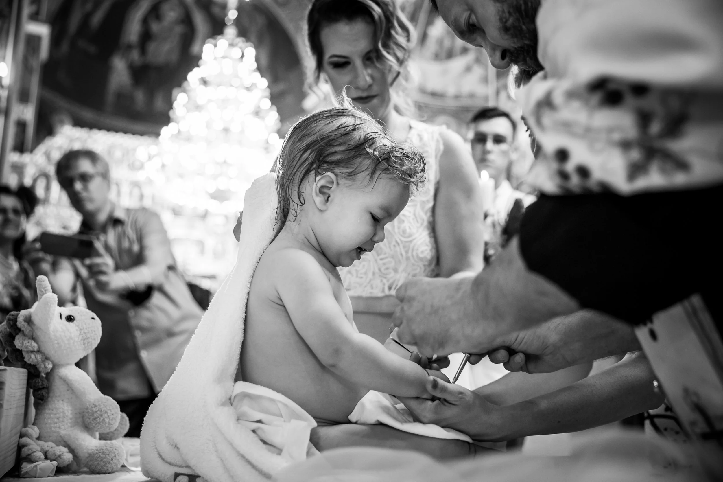 A young girl with wet hair and a towel around her shoulders receives a shot from a healthcare worker during a medical procedure, in a room decorated with blurred Christmas lights in the background, and an elf plush toy on the side.