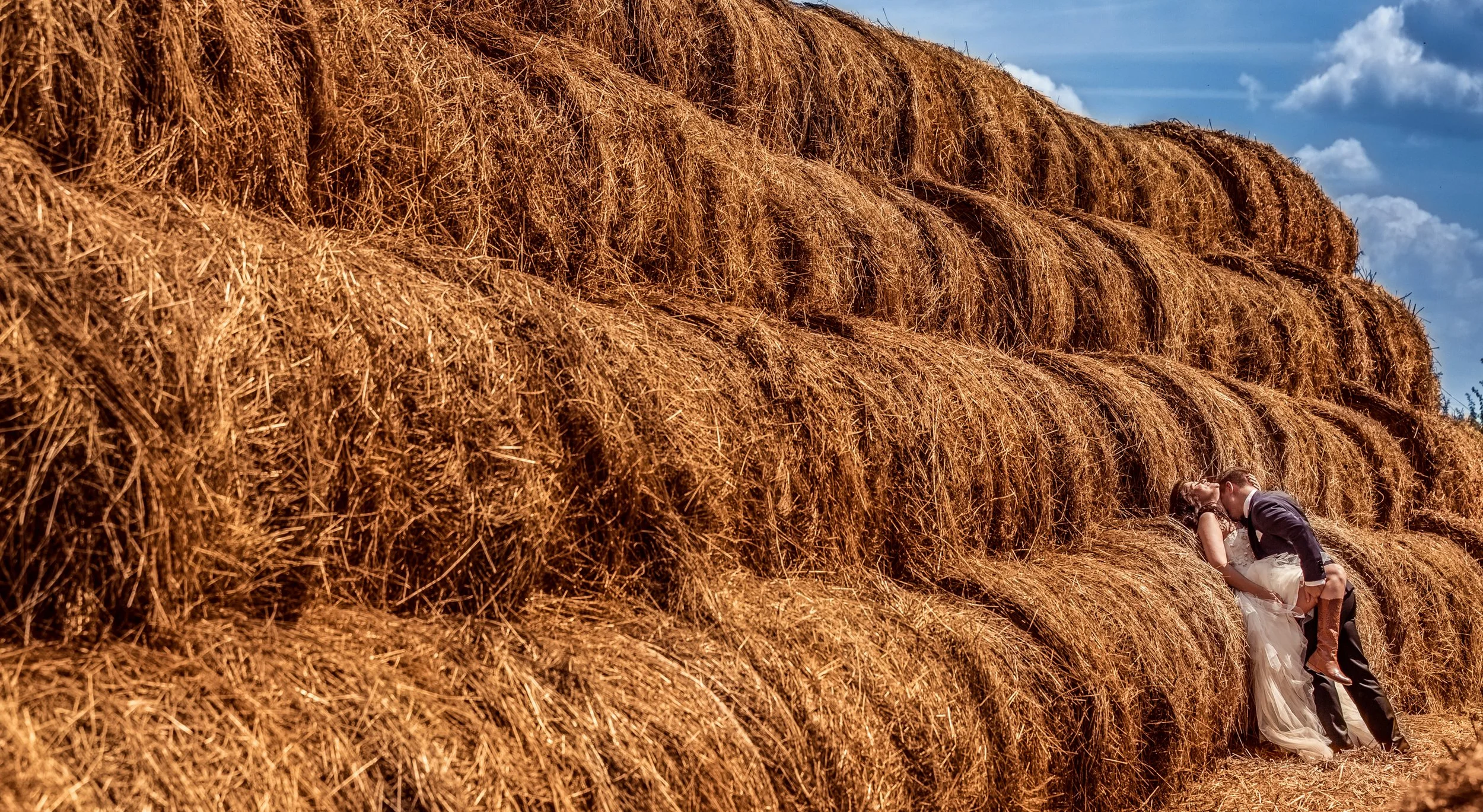 A bride and groom kiss in front of large hay bales under a blue sky with clouds.