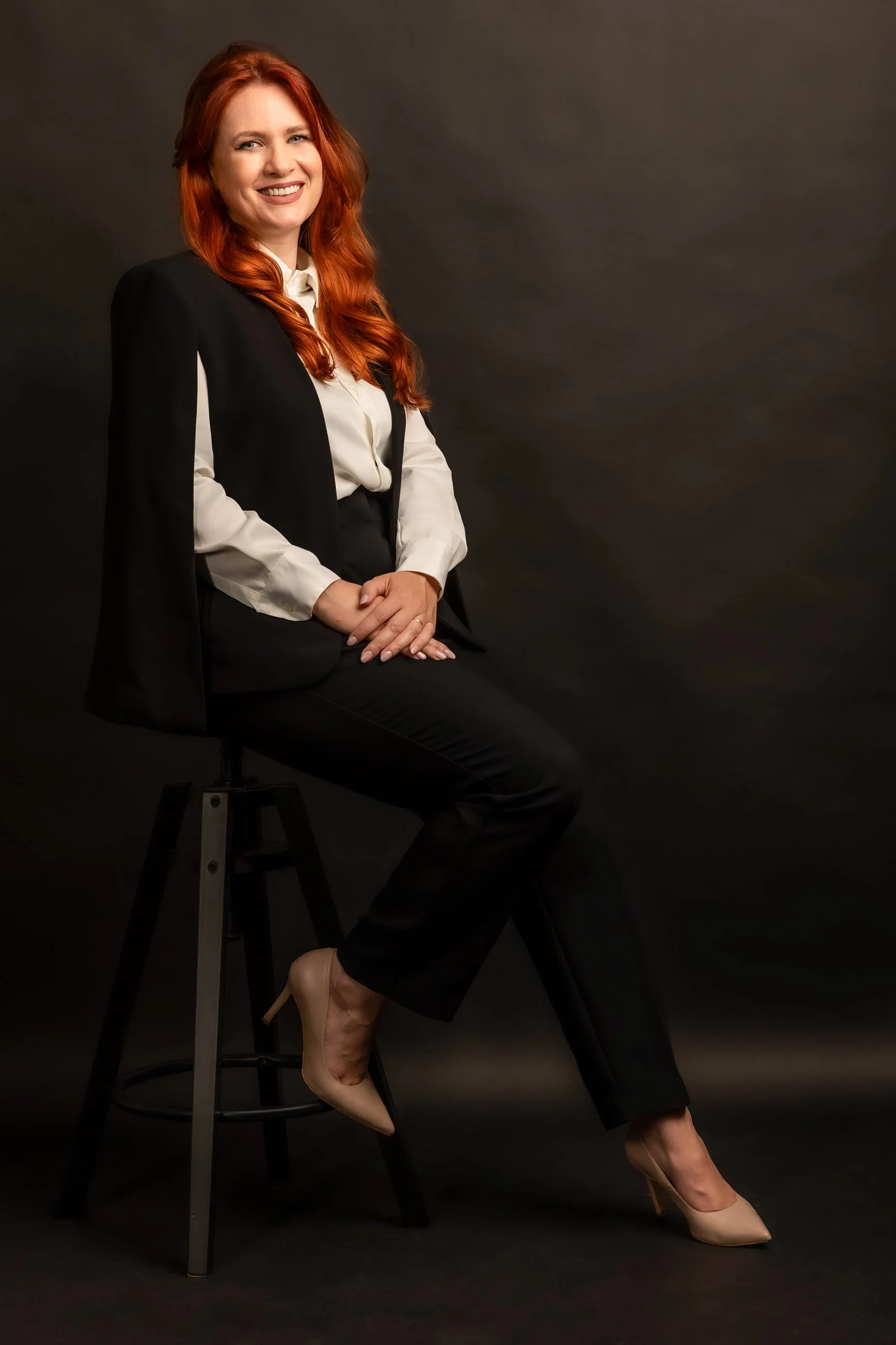 A woman with long red hair smiling while sitting on a black high stool against a dark background. She is dressed in a black blazer, white blouse, black pants, and beige heels, with her hands clasped in her lap.
