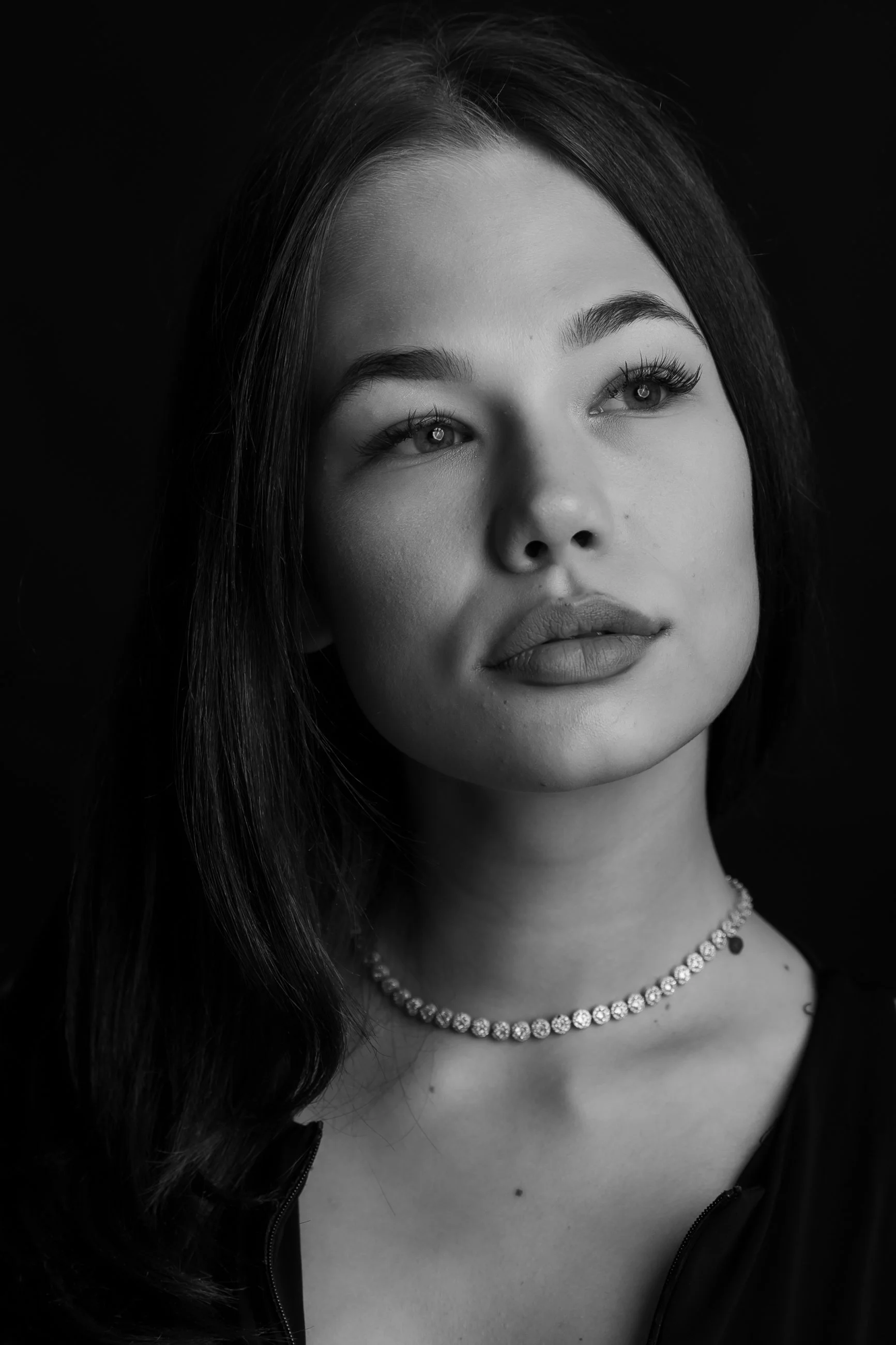 Close-up black and white portrait of a young woman with long hair, wearing a necklace with small stones and a partially unzipped top.