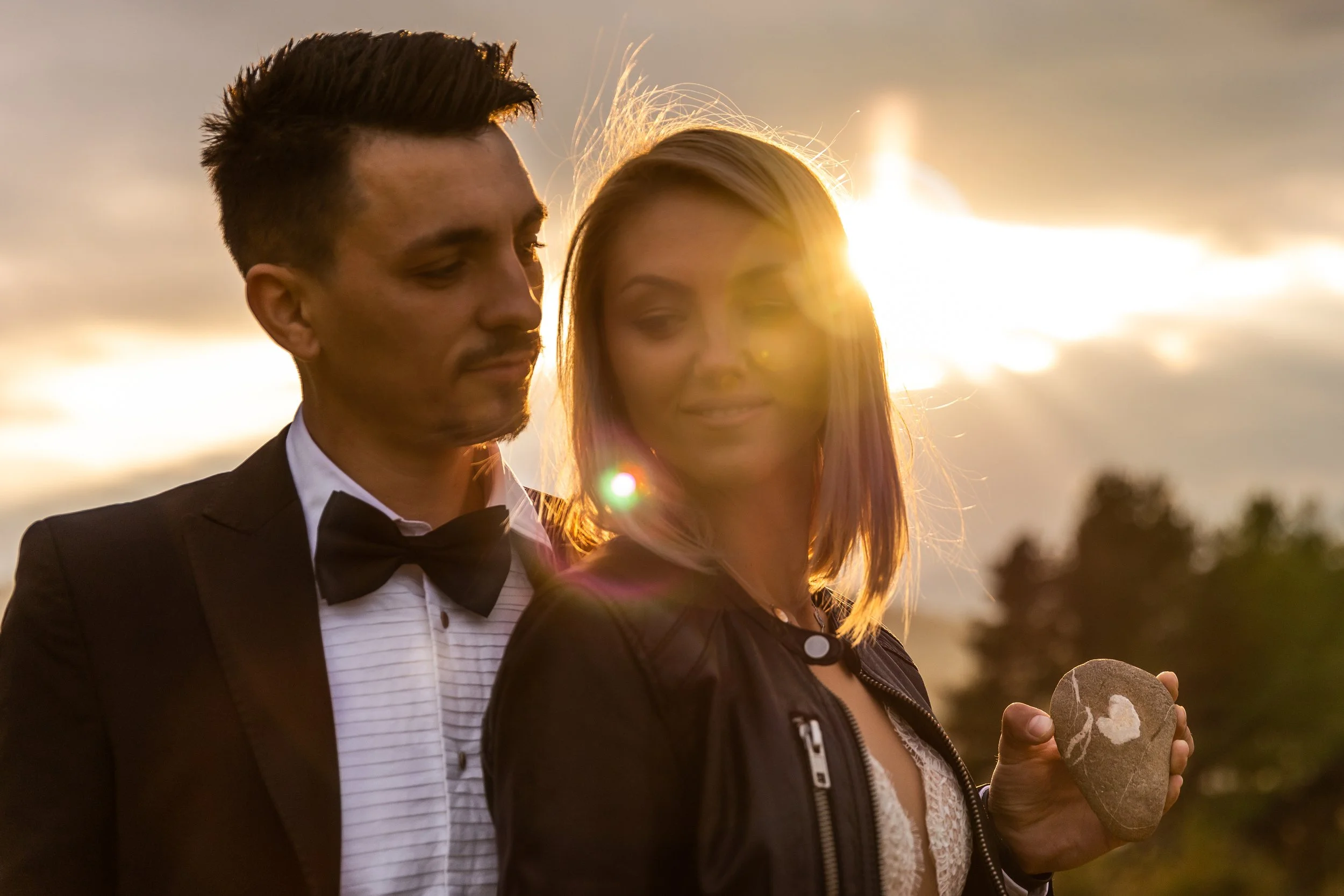 A young man in a tuxedo and a young woman in a dress standing outside during sunset, with the woman holding a painted rock.