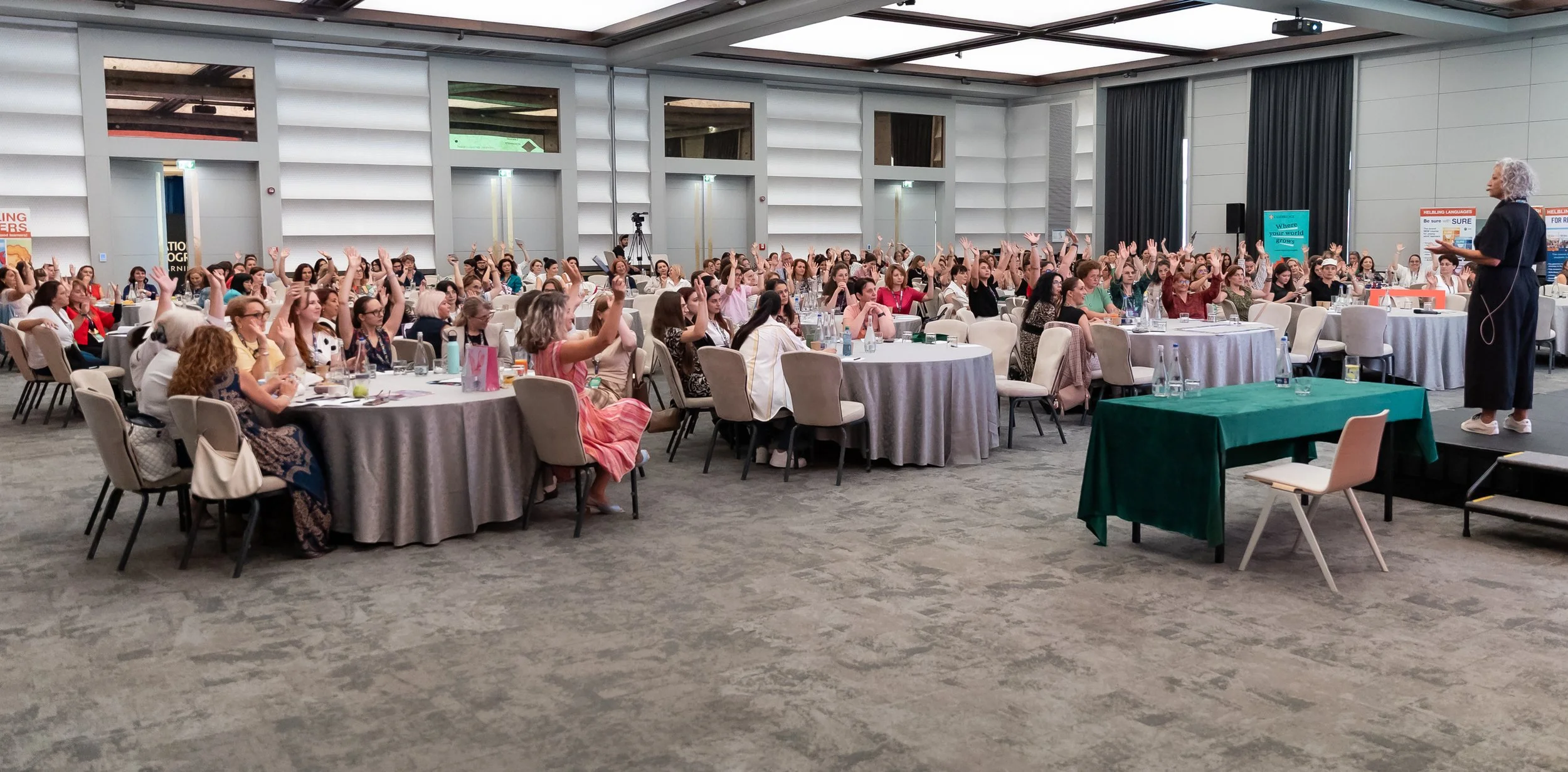 A large conference room filled with round tables and seated women attending a presentation, with a speaker standing at the front.