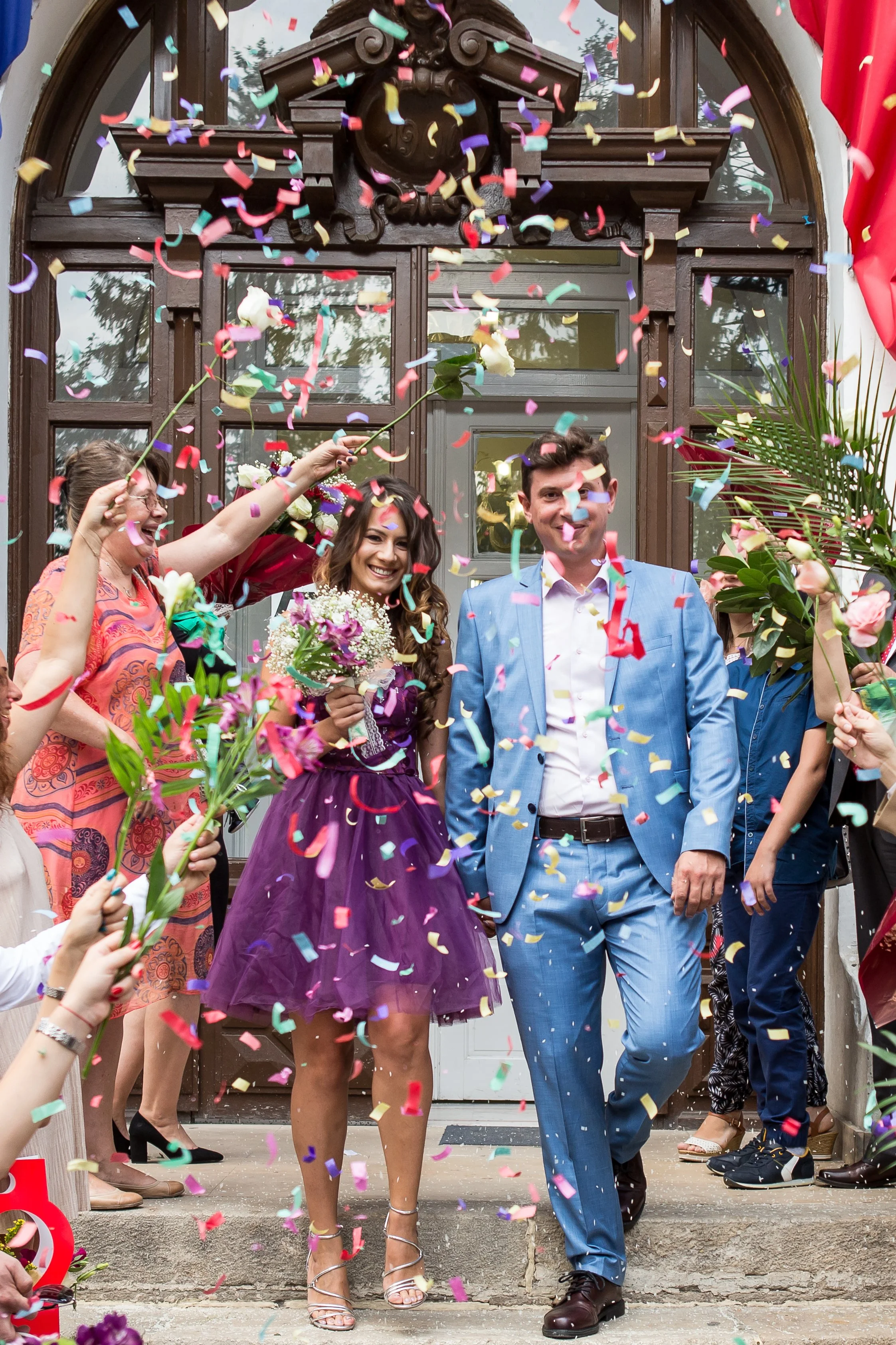 A wedding celebration with a bride and groom walking out of a building, surrounded by guests showering them with colorful confetti, flowers, and streamers, in front of a wooden door and windows.