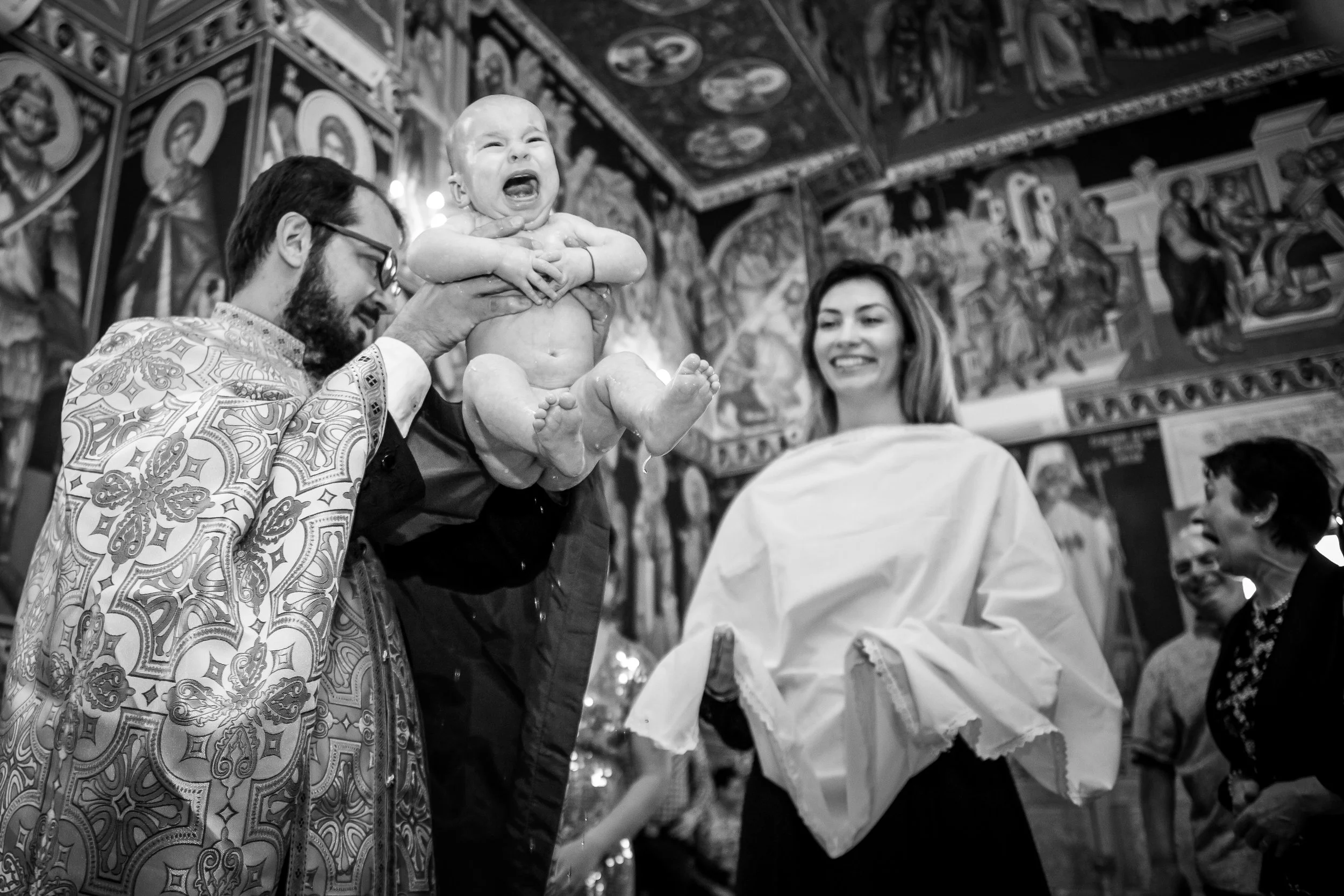 A baby being held up during a religious ceremony, crying with arms crossed, surrounded by smiling adults inside a church with ornate religious artwork on the walls.