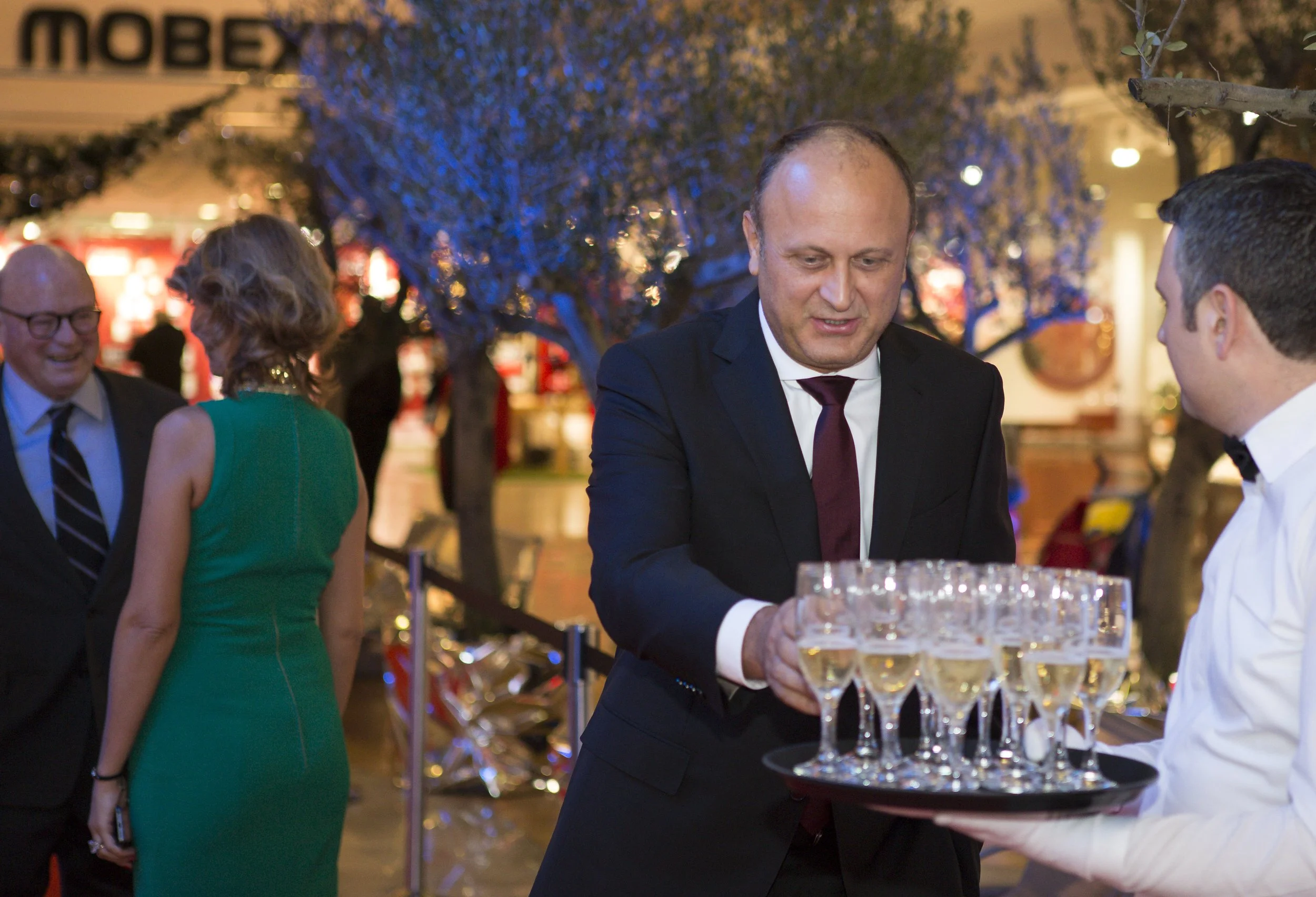 A man in a suit serving drinks at a bar during an indoor event. Other people are socializing in the background. The setting is decorated for an evening event with warm lighting and trees.