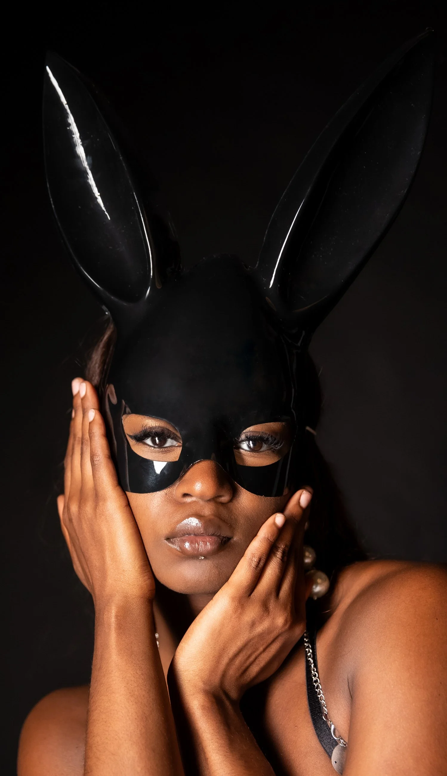A woman wearing a black bunny mask with tall ears, holding her face with both hands, against a black background.