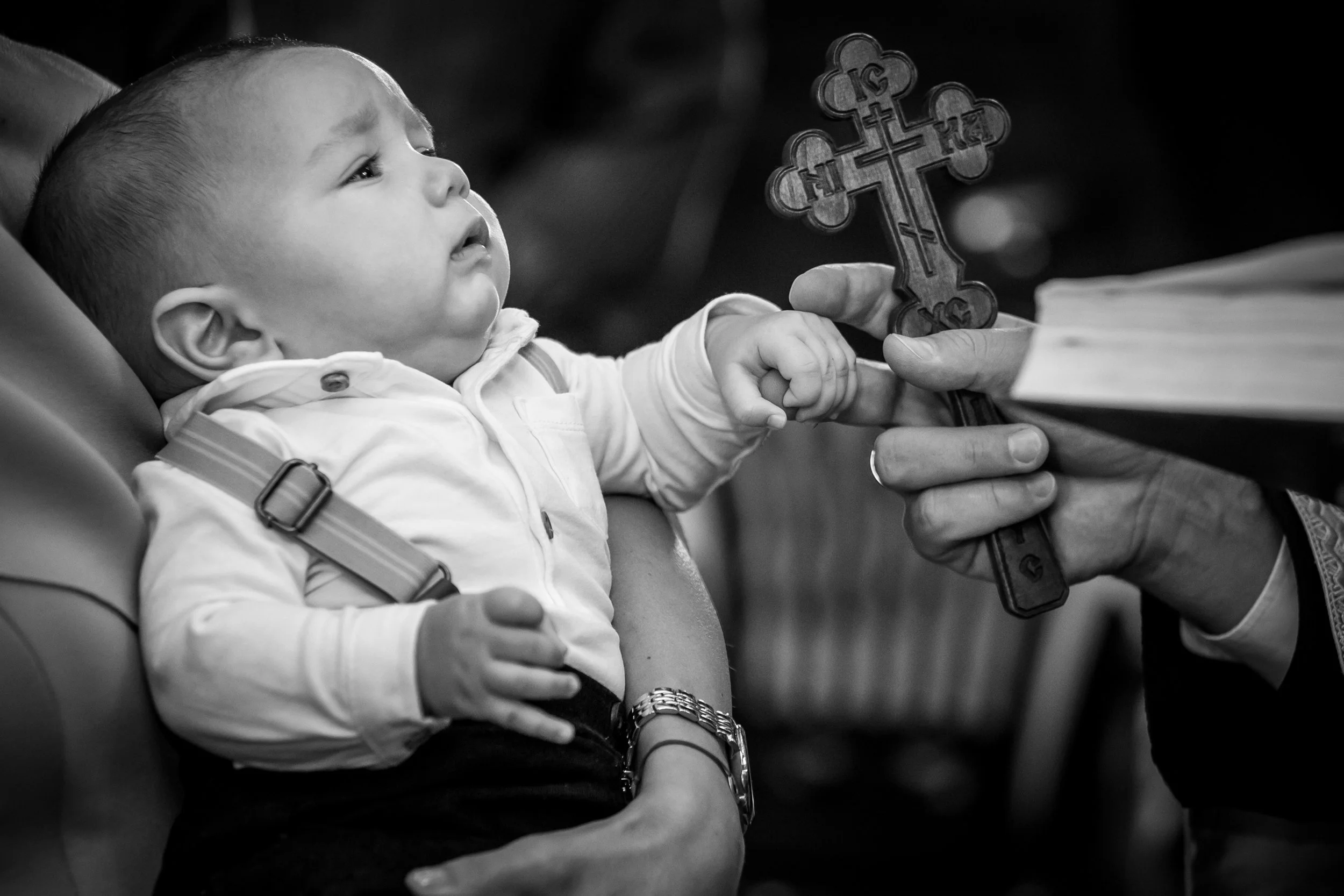 A young child with a halo of short hair sitting in a stroller, receiving a blessing or prayer from an adult. The adult, whose arm is visible, is holding a cross made of wood or metal, extending it toward the child's face. The scene appears to take pl